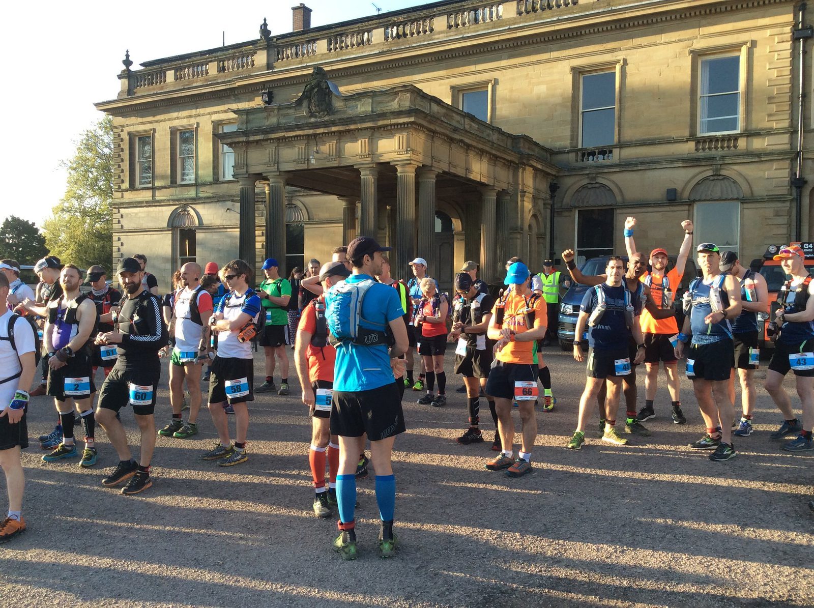 A group of runners gather in front of a large, historic building, preparing for a race. Some are stretching, while others chat or adjust their gear. They are wearing race bibs and athletic clothing with a variety of colors. The weather is clear and sunny.