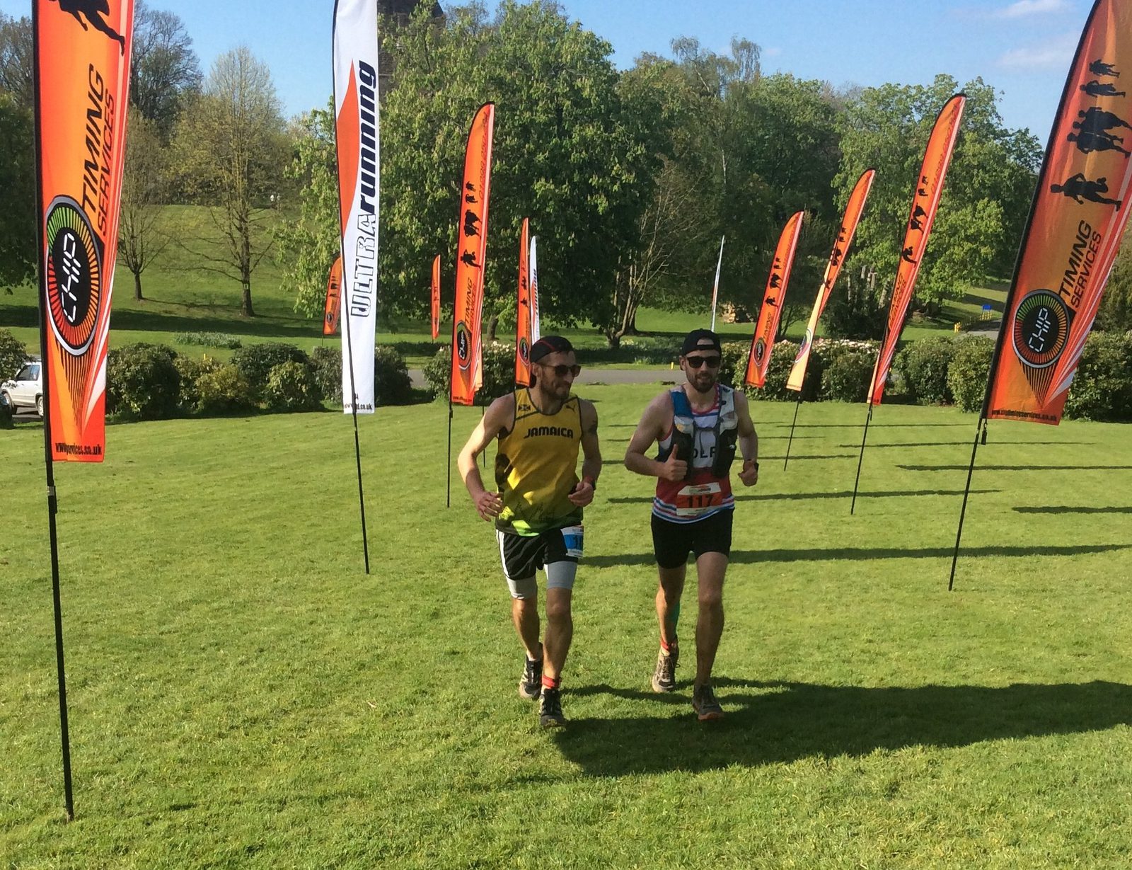 Two runners competing in a race on a grassy field, surrounded by orange and white flags bearing the text "CYD ULTRA RUNNING". The runners are in athletic gear under a clear sky and green trees in the background.