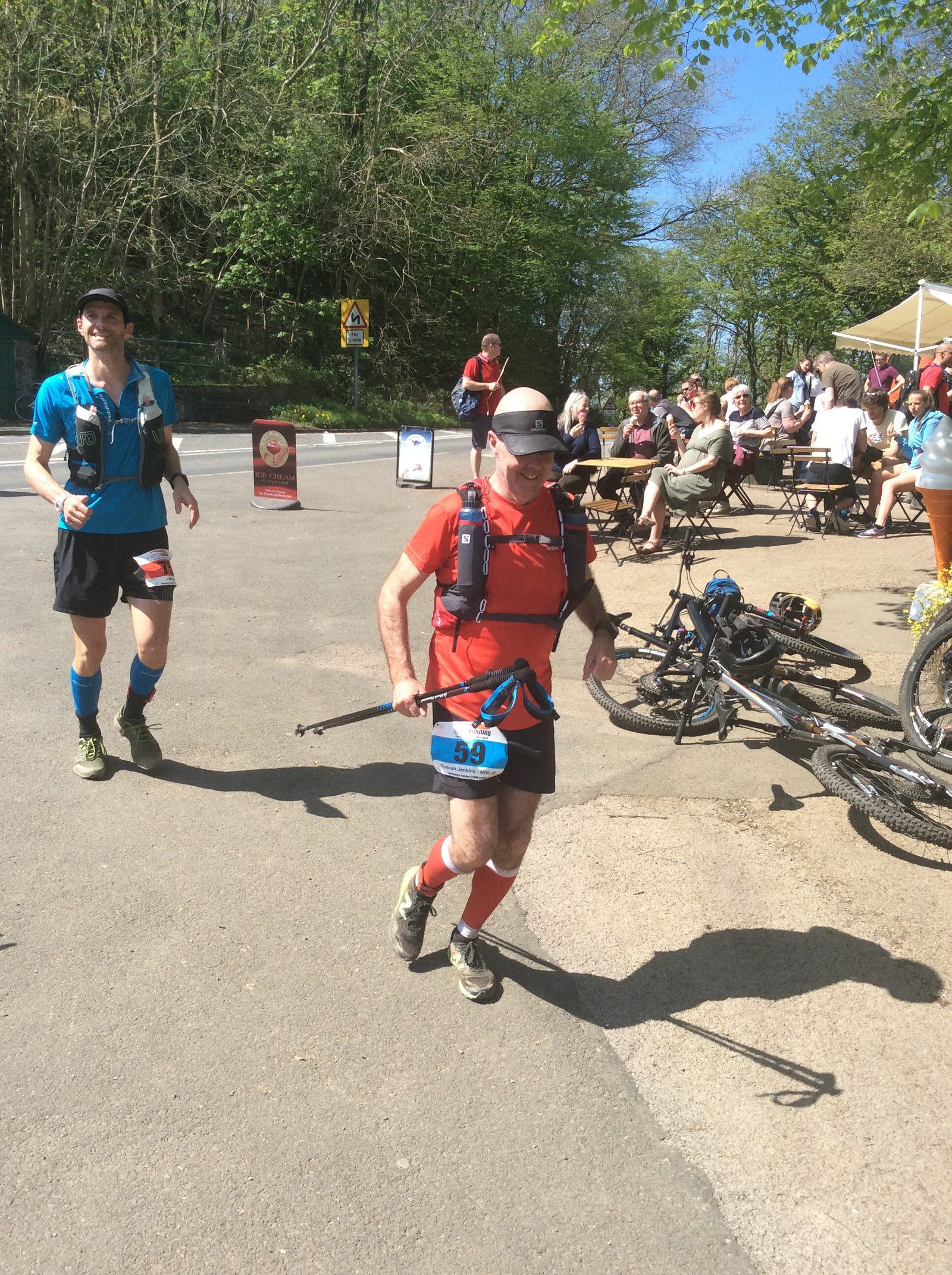 A man in a red shirt and black shorts runs with hiking poles in a sunny outdoor area, followed by another man in blue attire. Both wear race bibs and hydration packs. Spectators sit at picnic tables, and bicycles are parked beside them. Trees and signs are visible in the background.