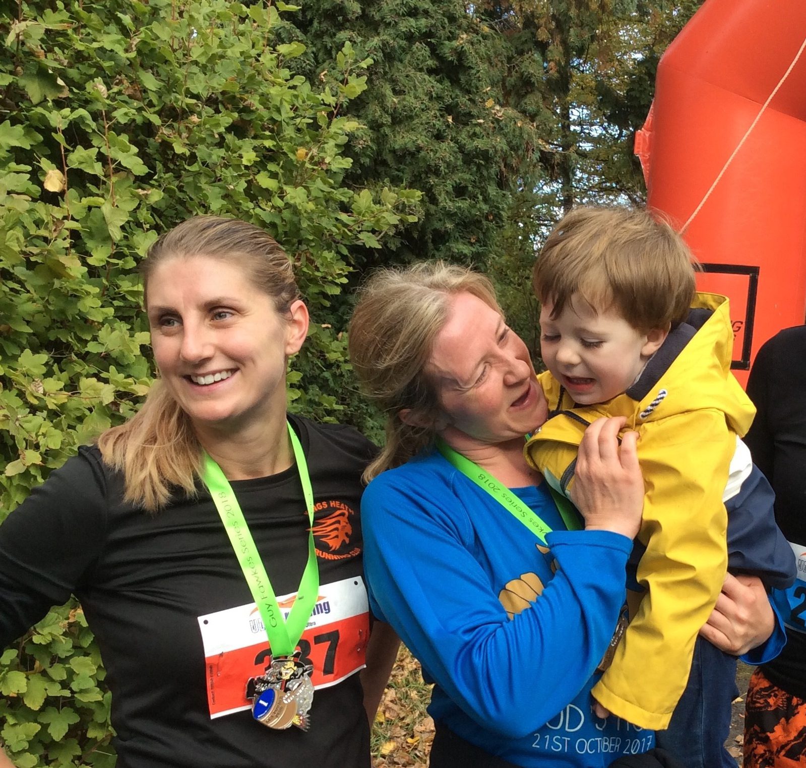 Two women with medals around their necks stand together outdoors, smiling and celebrating. The woman on the left wears a black shirt and has long, light brown hair. The woman on the right, in a blue shirt, is holding a young boy in a yellow jacket. Green foliage is in the background.