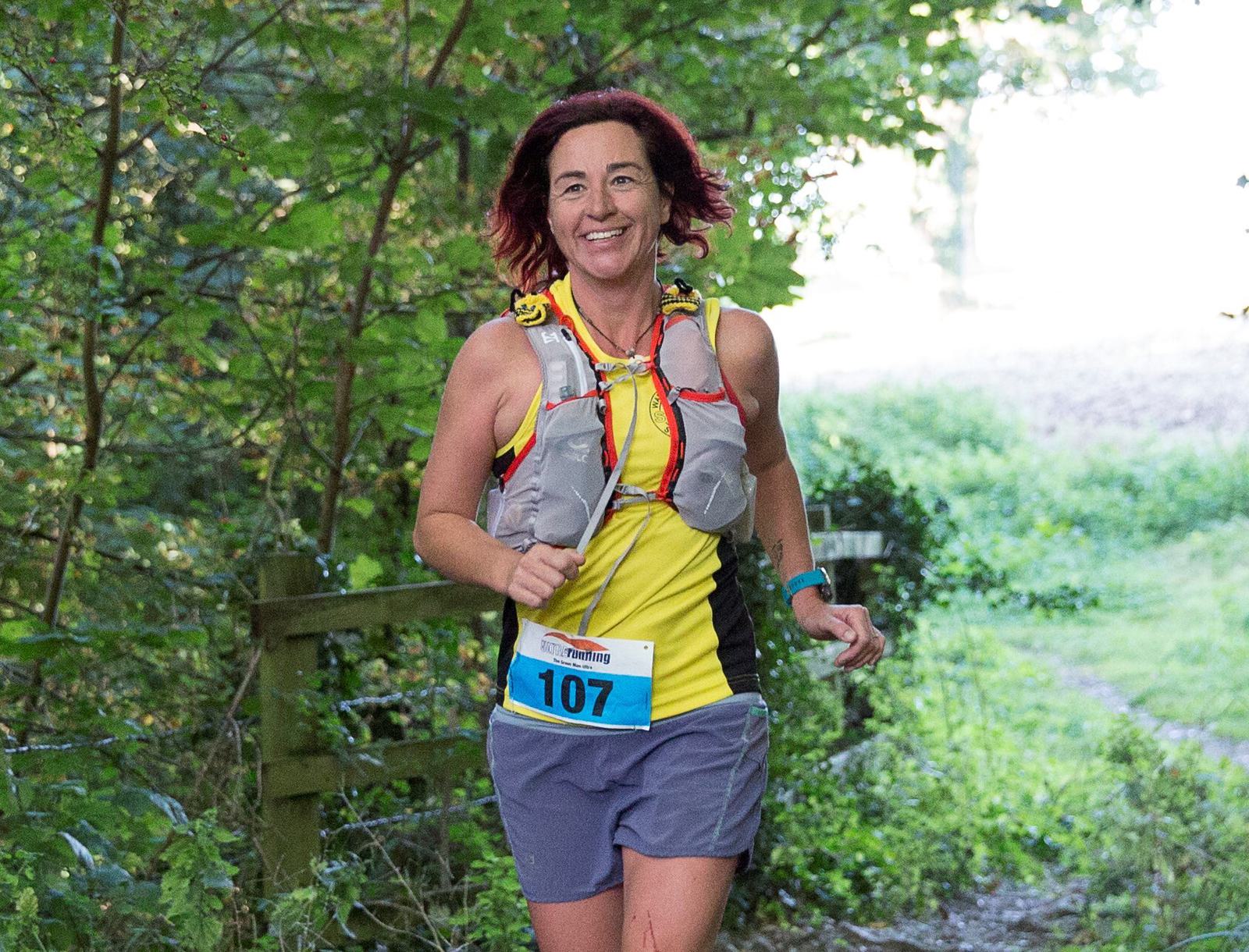 A woman with red hair smiles while running on a forest trail wearing a yellow shirt, gray shorts, and a hydration vest. She has a race number 107 bib pinned to her shorts. Surrounding her are lush green trees and foliage.