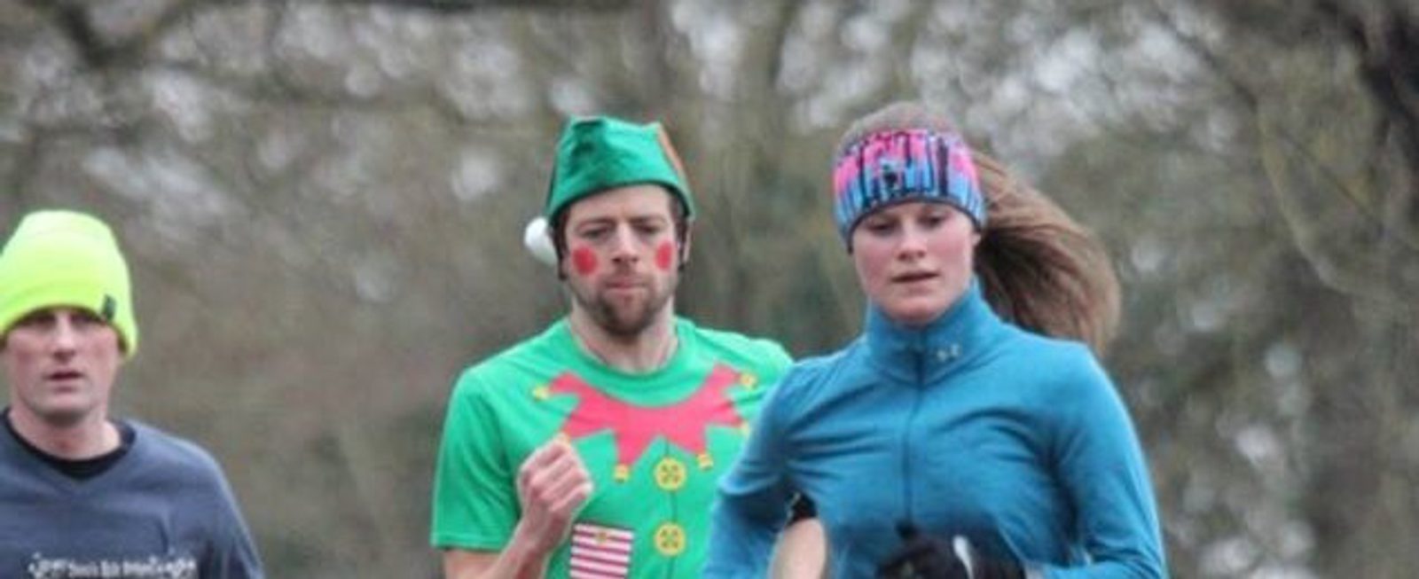 Three people are running outdoors on a cloudy day. The person in the center is wearing a green elf costume with red cheeks, the person on the right is dressed in a blue jacket and a colorful headband, and the person on the left is wearing a neon yellow hat and a dark shirt.