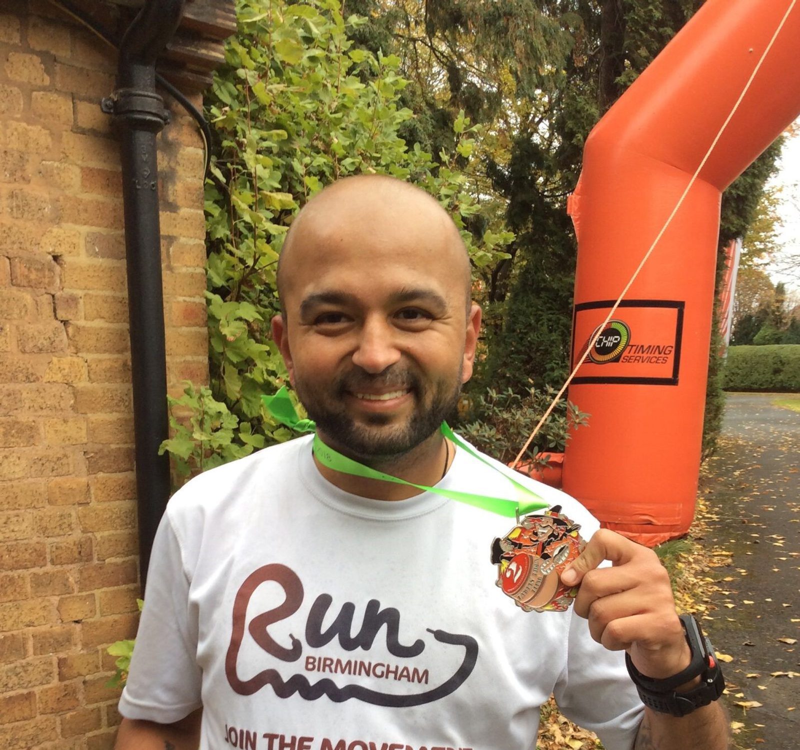 A man with a shaved head and beard smiles while holding a medal and wearing a white "Run Birmingham" shirt. He is standing outdoors next to a brick building and an orange inflatable archway. Lush green foliage is visible in the background.