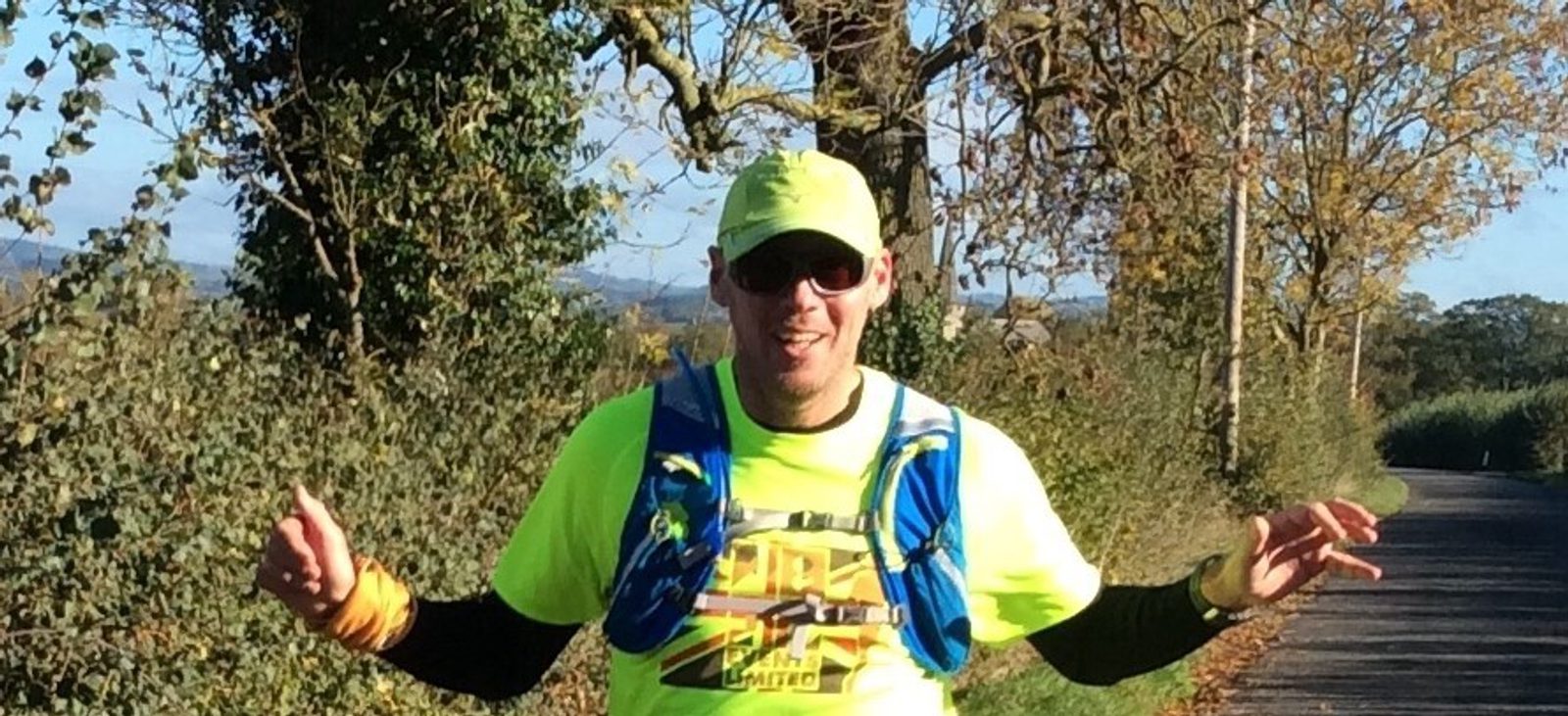 A person wearing a bright neon yellow shirt, cap, and a hydration pack is running along a rural road lined with trees and shrubs. They are smiling and have their arms raised as if waving or gesturing. The weather is sunny with a clear sky in the background.