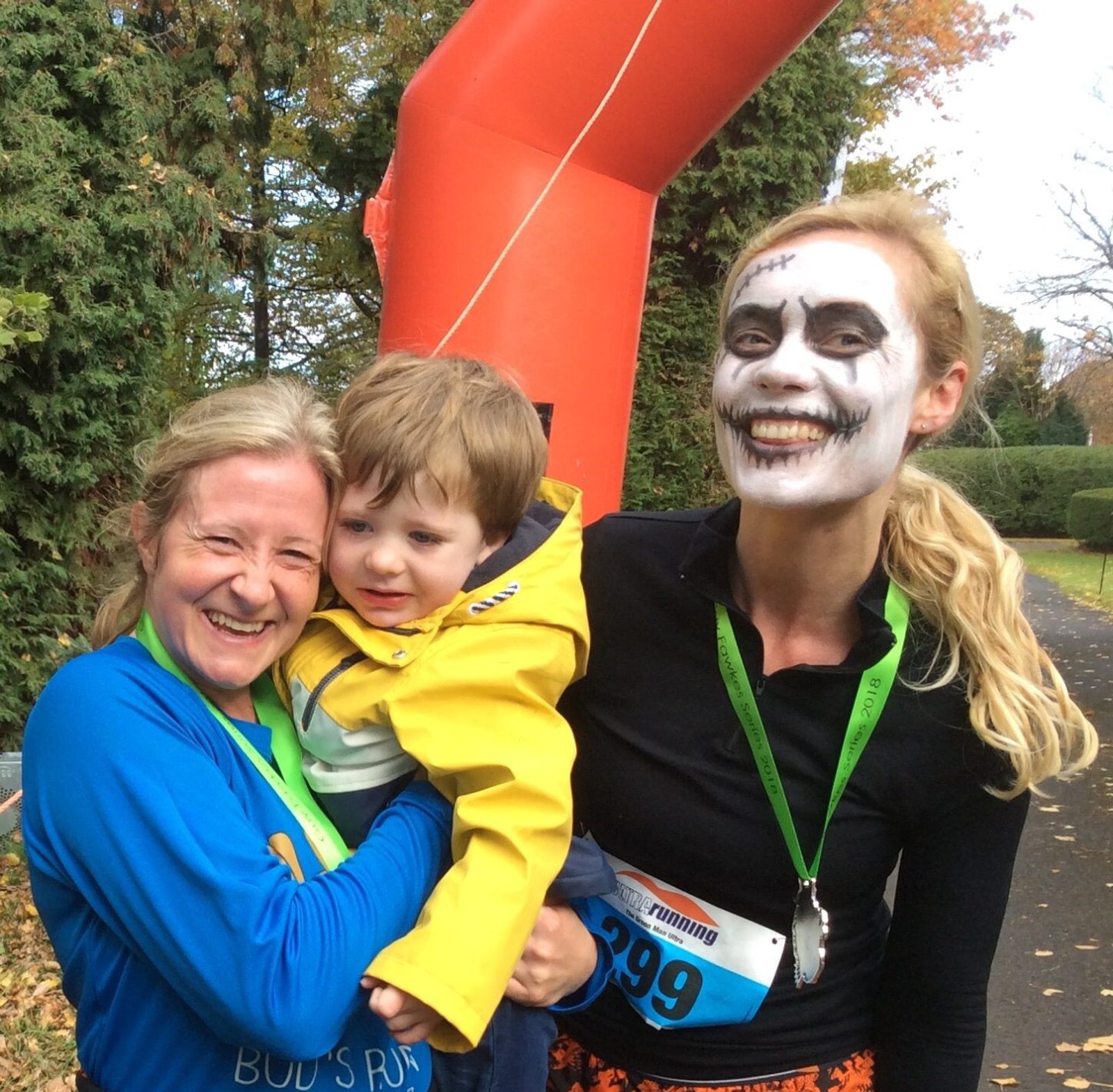 Two women and a child are posing and smiling. One woman is in a blue shirt and holds the child wearing a yellow jacket. The other woman is wearing a skeleton face paint, black shirt, and race bib number 299. They are outdoors with trees and an orange inflatable structure in the background.