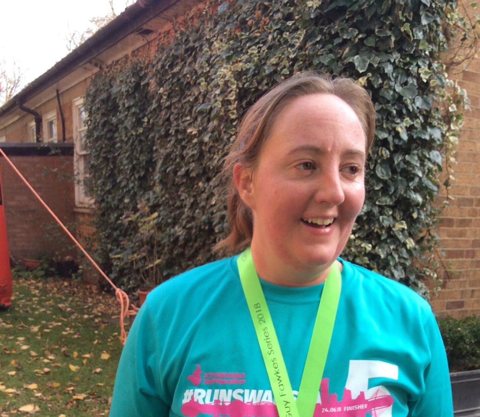 A person smiles while standing outside near a hedge. They are wearing a teal T-shirt with pink and white text and a neon green lanyard. The background includes shrubs and a brick building. The atmosphere appears to be relaxed and joyful.