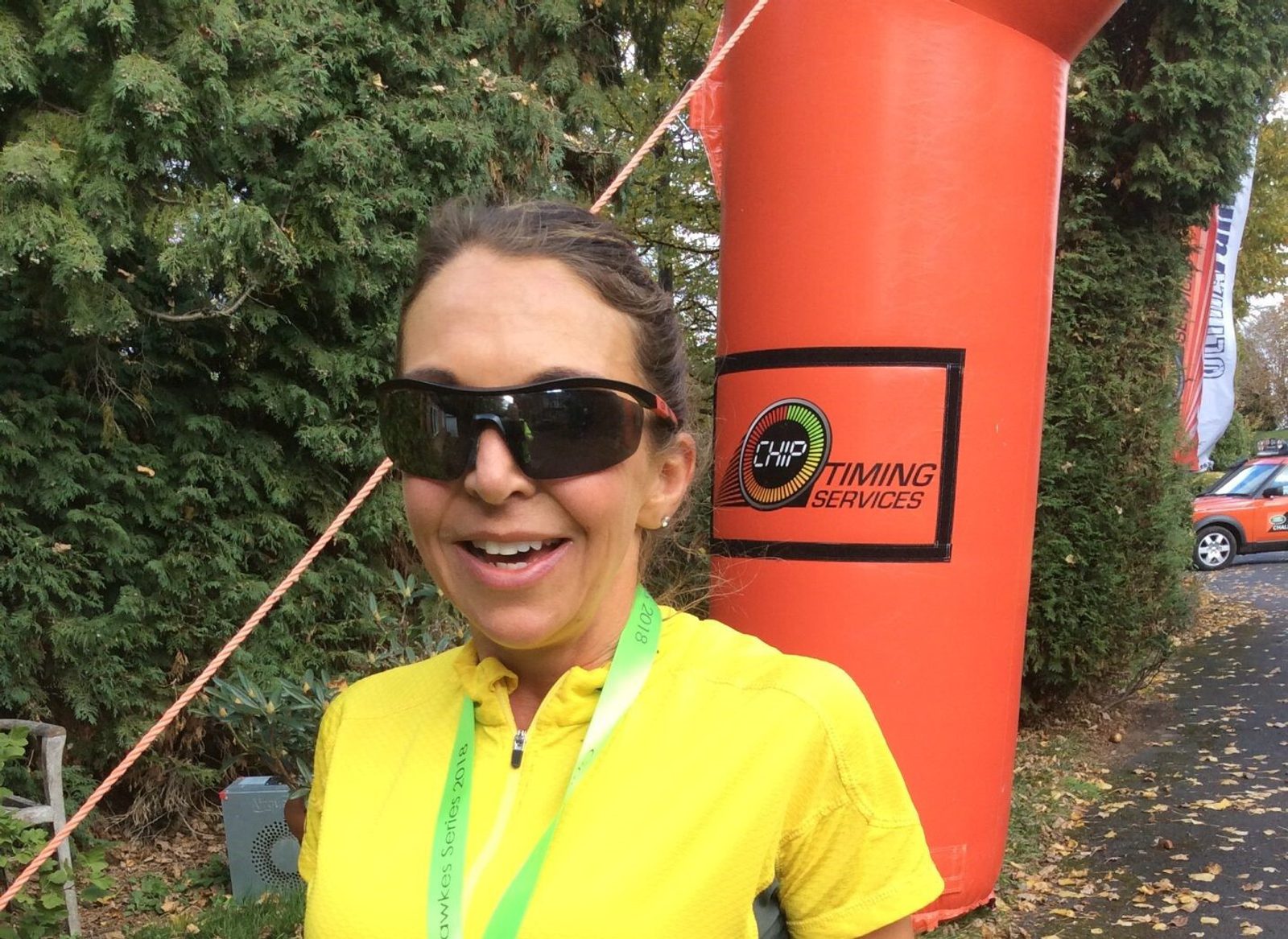 A person wearing dark sunglasses and a bright yellow shirt smiles at a race event. They have a green medal around their neck, and an orange inflatable marker with "Timing Services" is visible in the background. Trees and some fallen leaves are also in the scene.