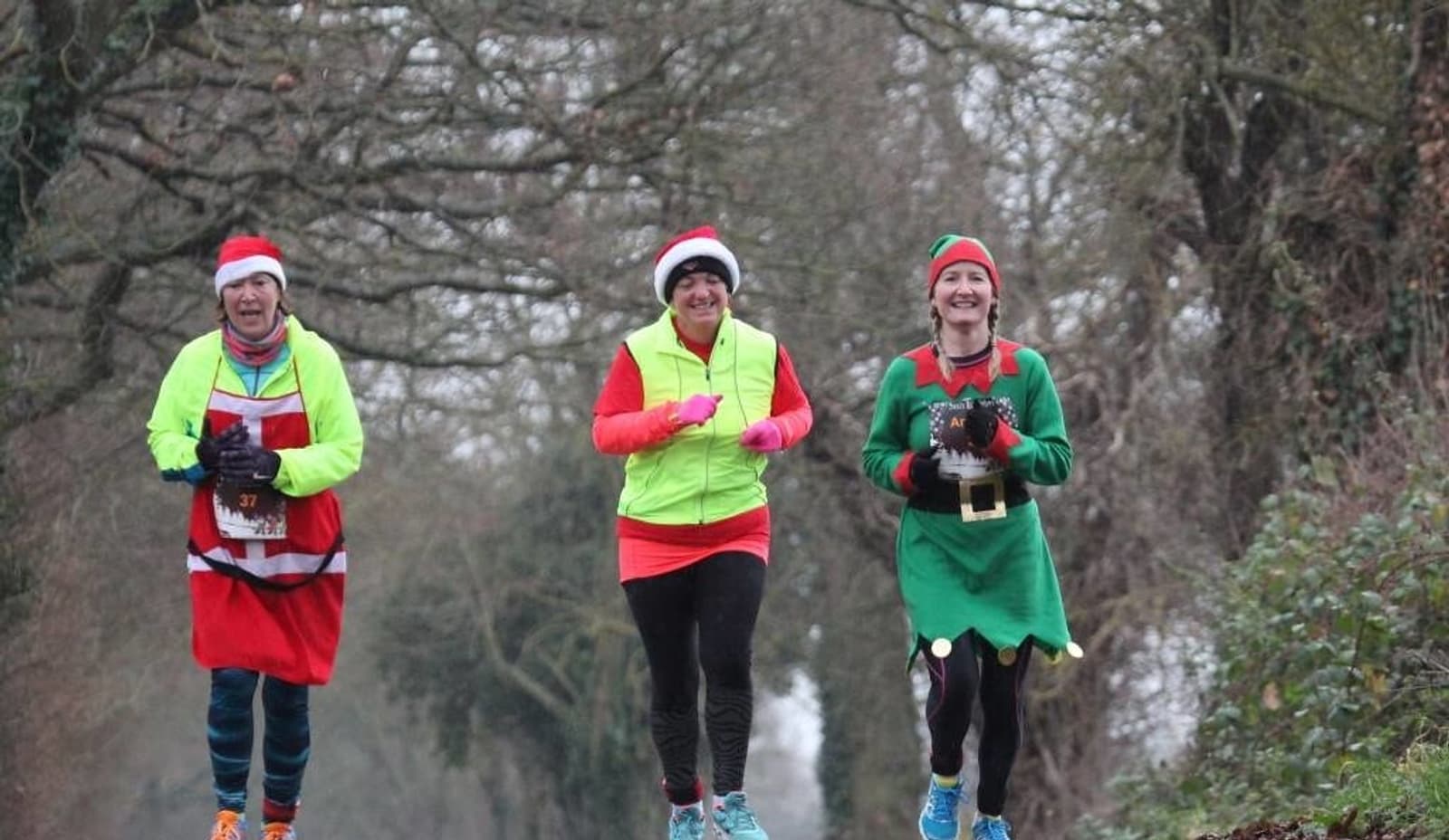 Three people are jogging outdoors on a wooded path wearing festive costumes. One is dressed as Santa, another in a Santa hat and neon yellow vest, and the third as an elf. The scene appears to be a winter or holiday-themed run.