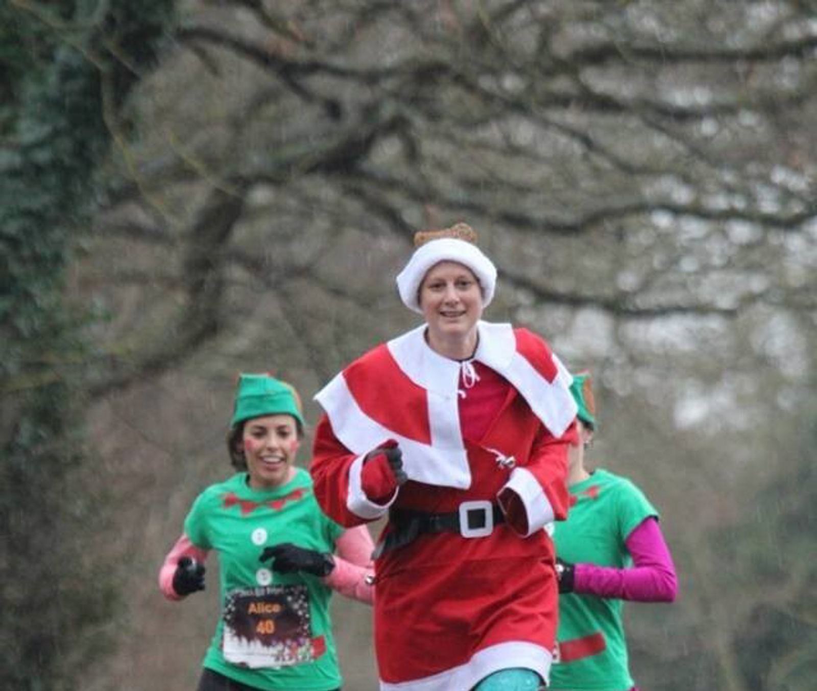 A person dressed in a Santa Claus costume is running outdoors, accompanied by two people in green elf outfits. Trees with bare branches are visible in the background, suggesting a winter setting. The runners are smiling and appear to be in a festive, joyful mood.