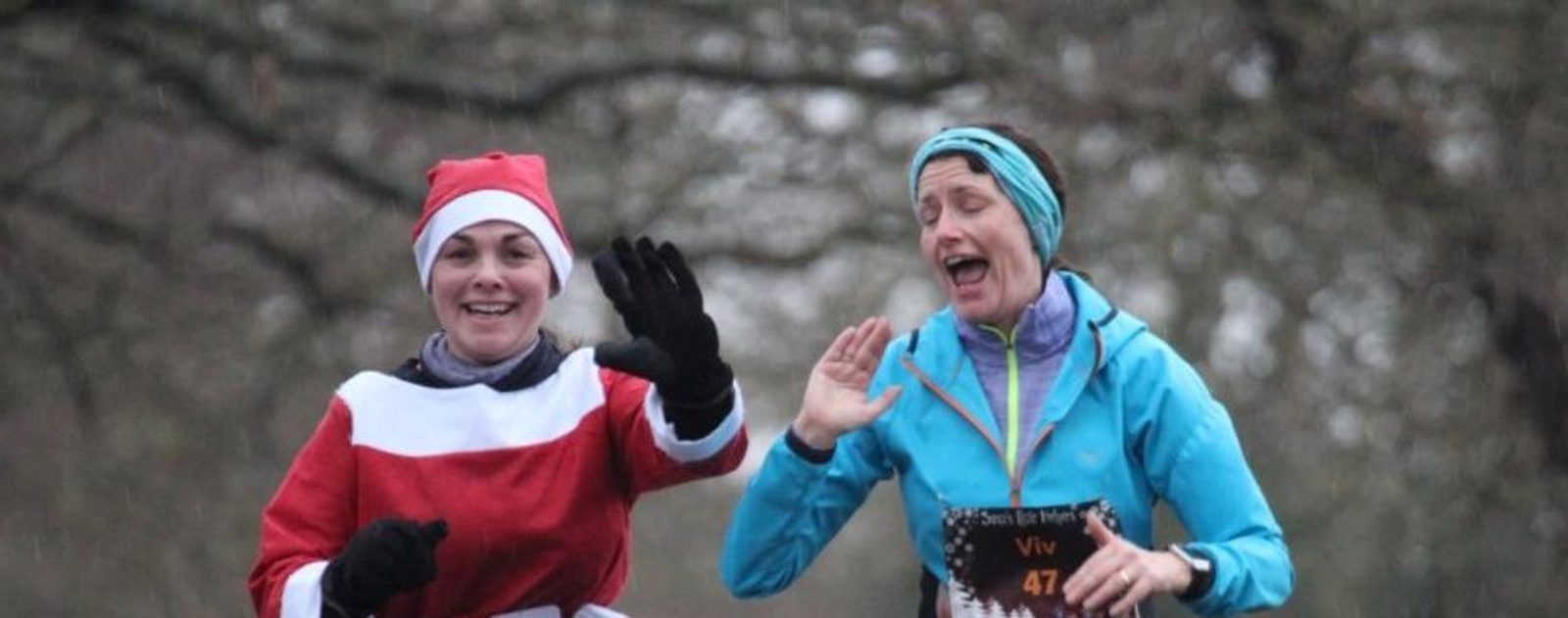Two people are running outdoors. One is wearing a Santa costume and the other is dressed in a blue jacket with a headband. Both appear to be joyful, smiling and waving as they move along the path. Trees and overcast skies form the background.