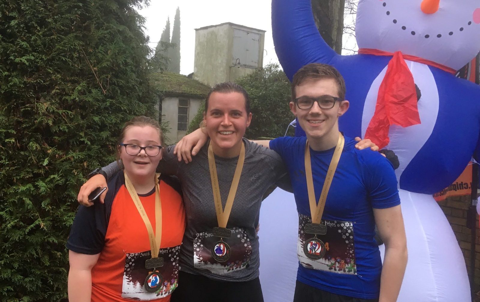 Three smiling people wearing medals stand with their arms around each other in front of an inflatable snowman. The two individuals on the left wear sports shirts, while the person on the right wears a blue shirt. Trees and a building are visible in the background.