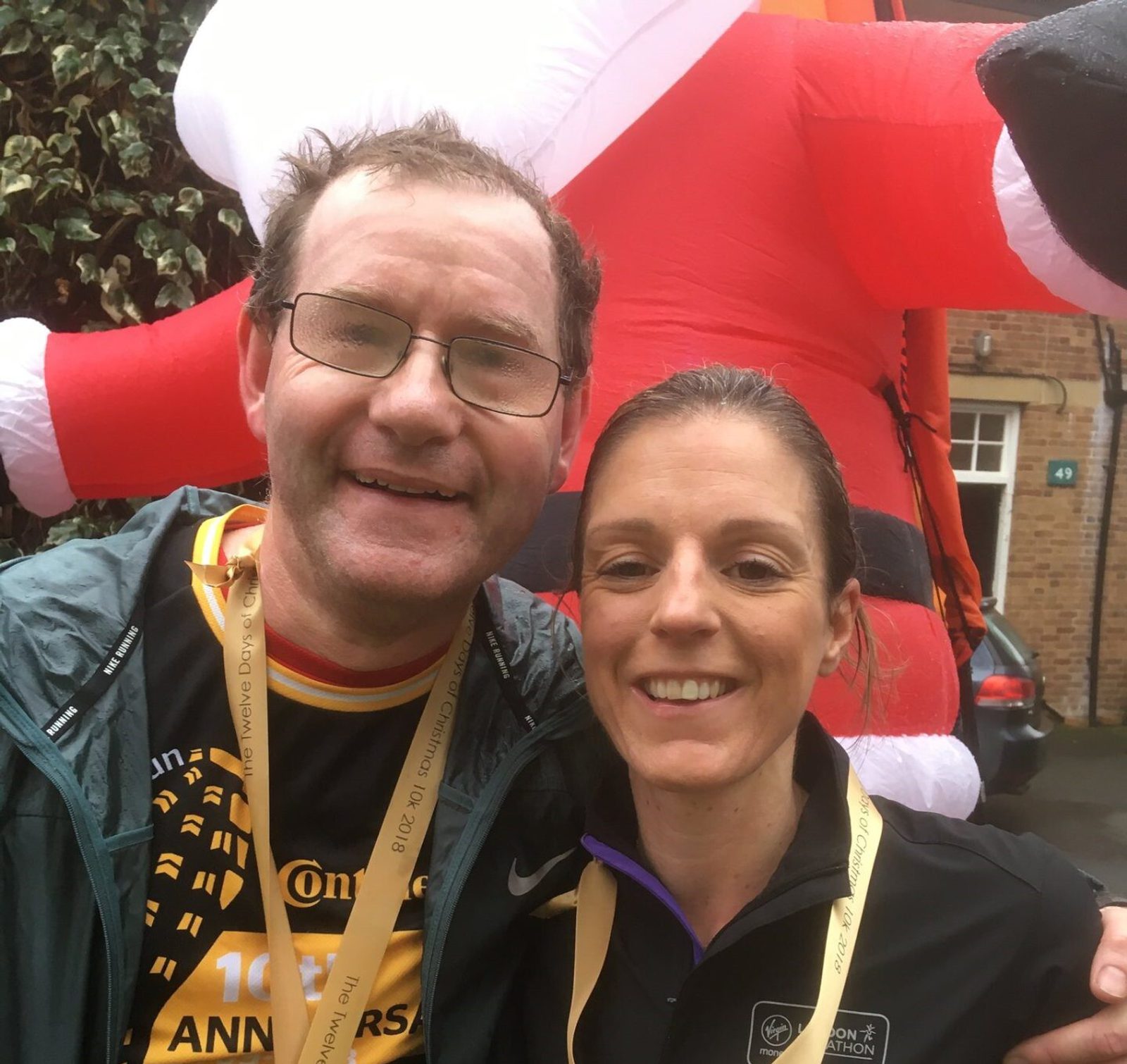 Two people, a man and a woman, smile at the camera with an inflatable Santa Claus in the background. Both wear medals around their necks, suggesting they completed a race or event. The man has glasses and short hair, while the woman has her hair pulled back.
