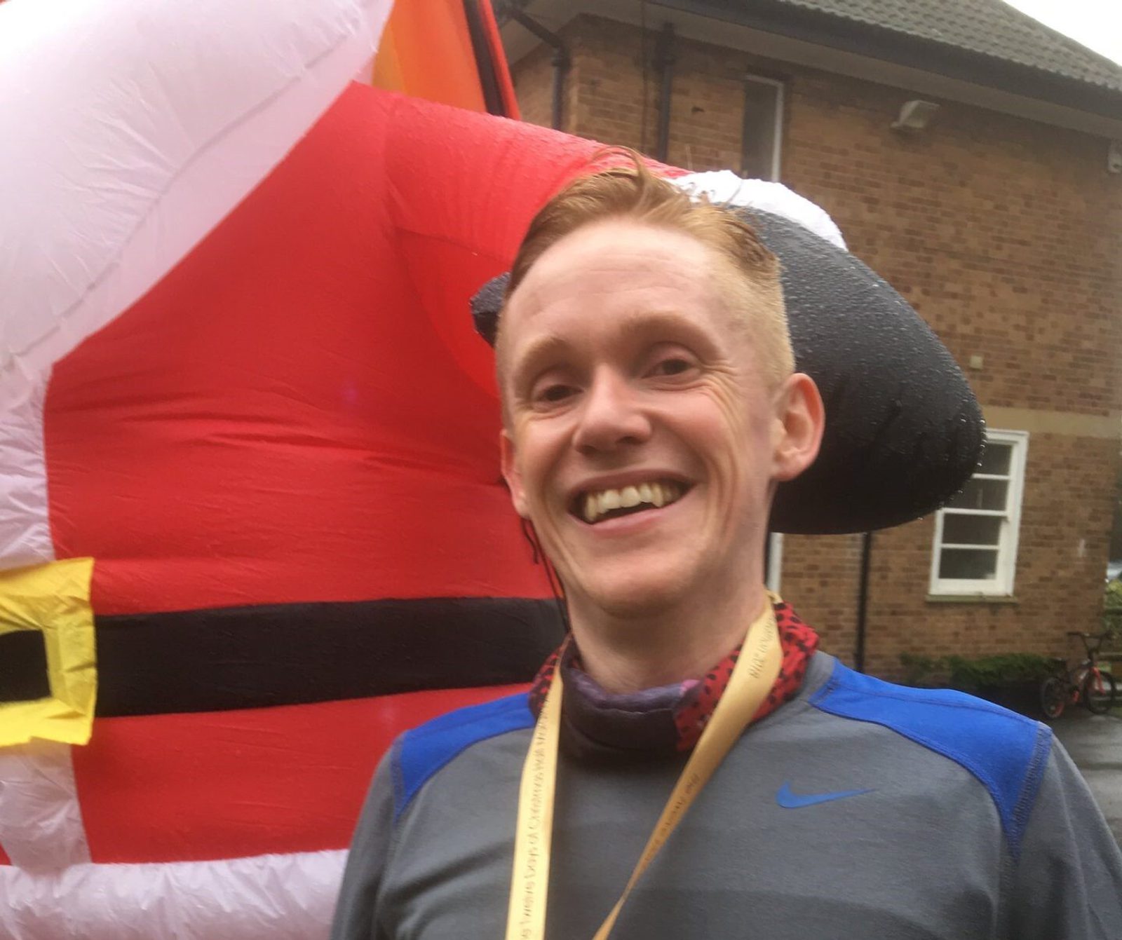 A person with a smiling expression stands in front of an inflatable Santa Claus. They are wearing a grey and blue sports outfit with a medal hanging around their neck. There's a building with brick walls and windows in the background.