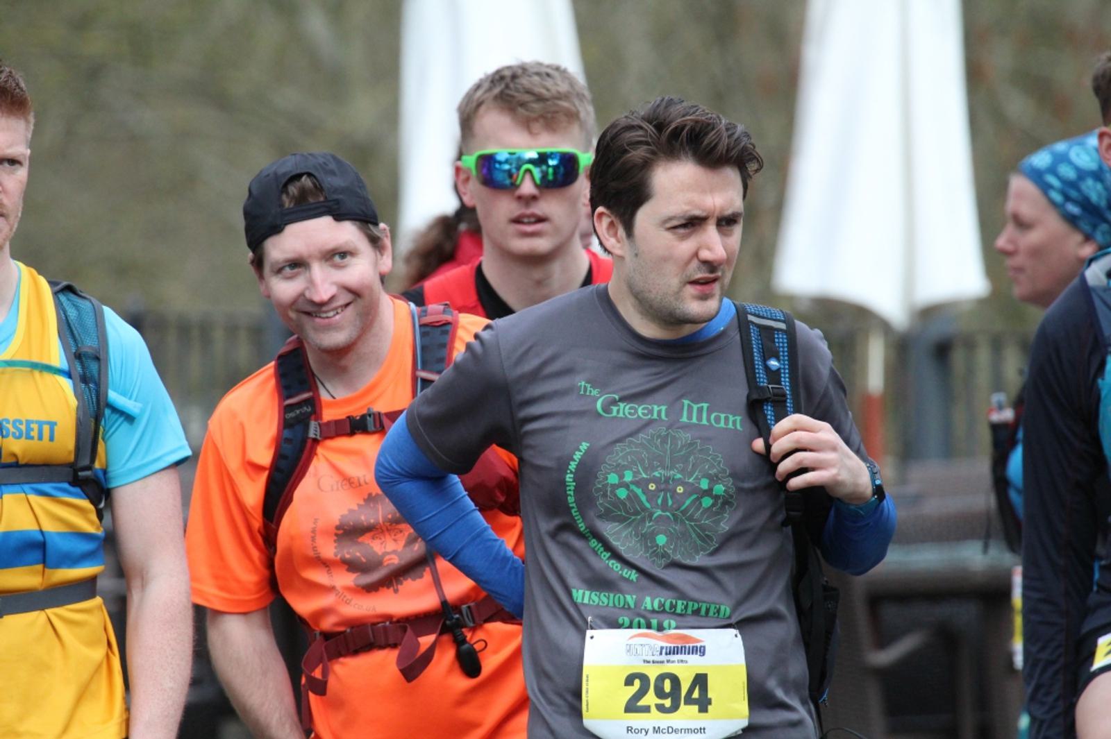 A group of runners, wearing race bibs and backpacks, stand together outdoors. One runner in an orange shirt and black cap smiles at the camera. Another in a gray shirt and yellow bib, marked 294, looks ahead thoughtfully. Other participants are visible in the background.