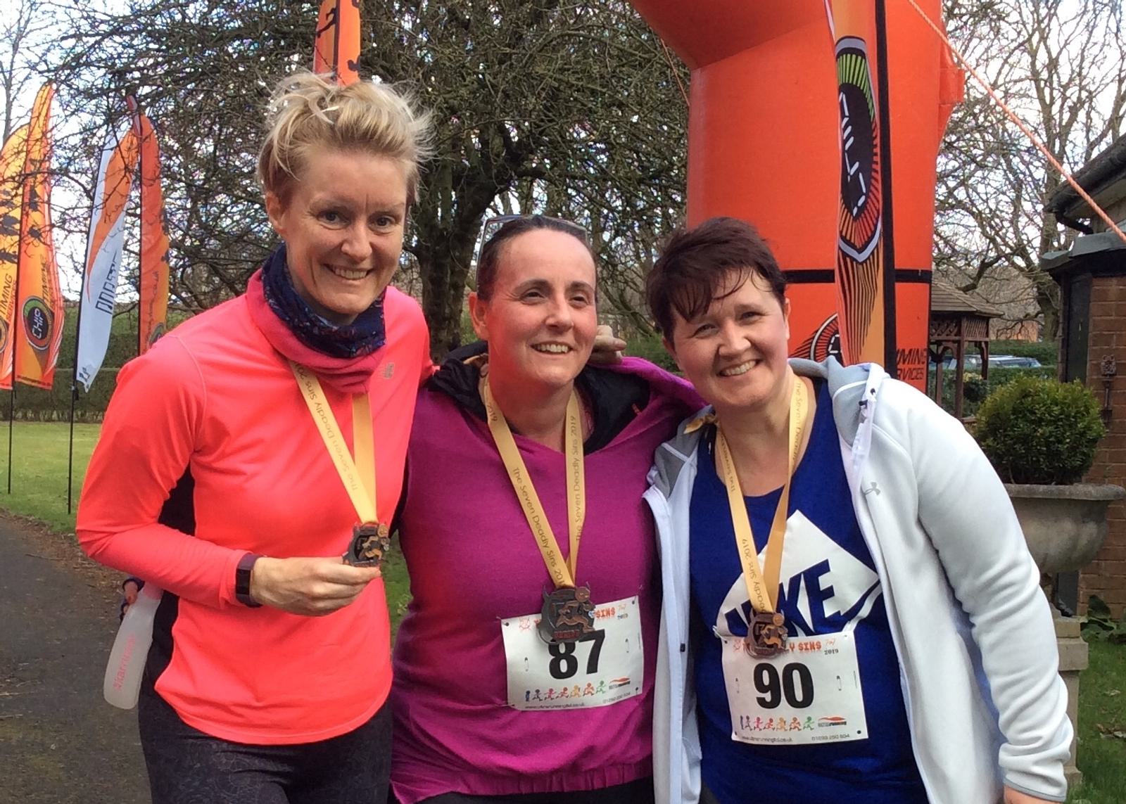 Three women, each wearing a medal around their neck, pose together after a race. They are smiling and appear happy. The woman on the left is in a bright pink top, the middle woman is in purple with race number 87, and the woman on the right in a white jacket with race number 90. They stand in front of race banners and an inflatable archway.