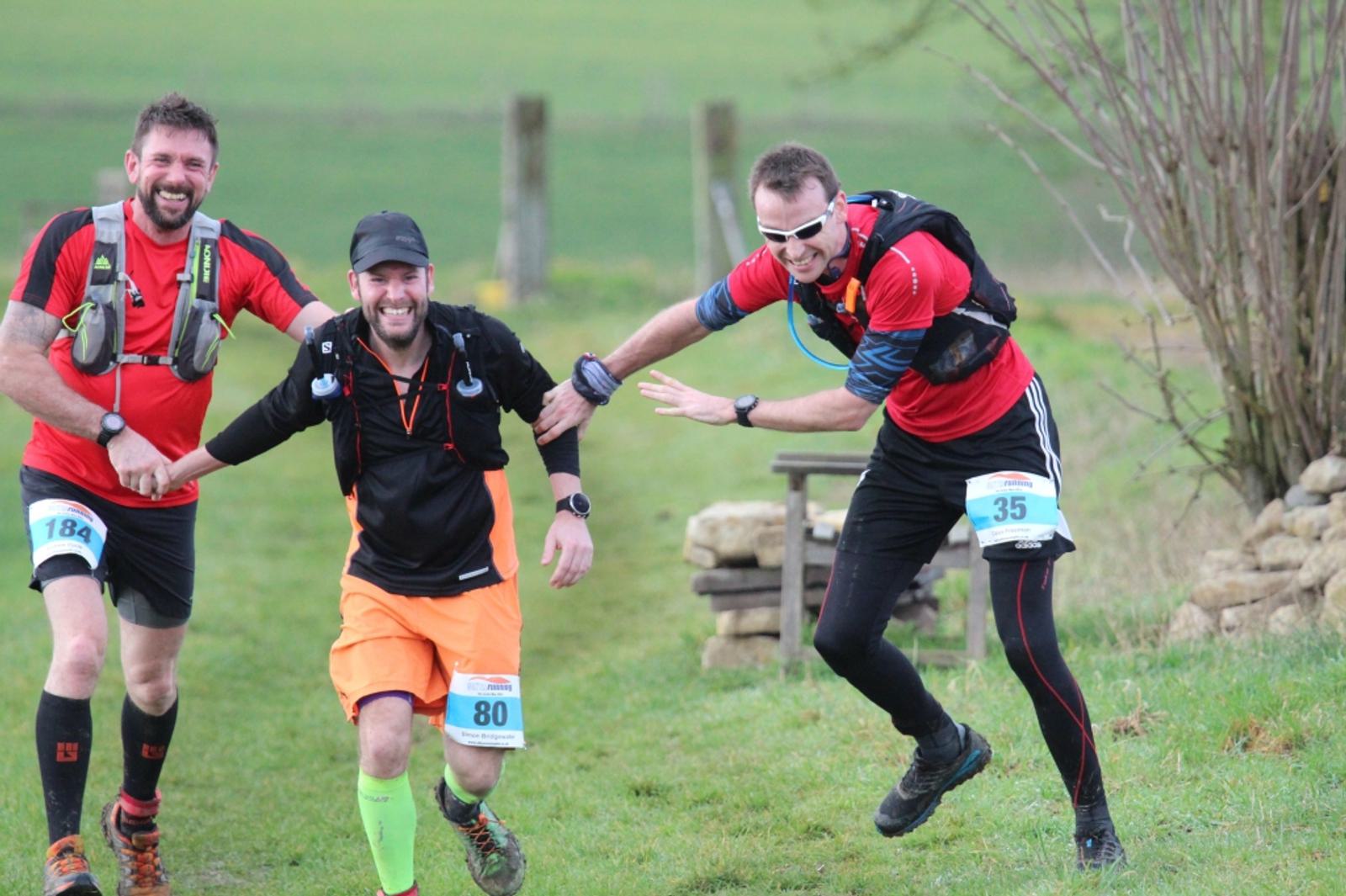 Three male runners, all wearing race bibs and sports gear, are running joyfully outdoors on a grassy path. One of them is playfully holding another’s hand. They appear to be enjoying the run together, with all of them smiling. The background features a green field.