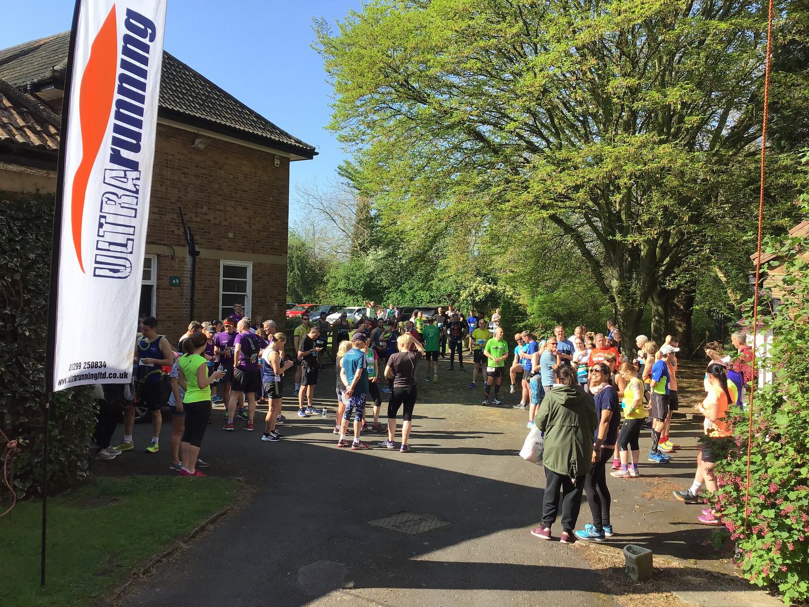 Runners gather outside near a brick building and trees on a sunny day, preparing for a race. A white flag with "Ultra Running" text stands on the left. Participants are dressed in colorful athletic gear, with some stretching and others chatting in groups.