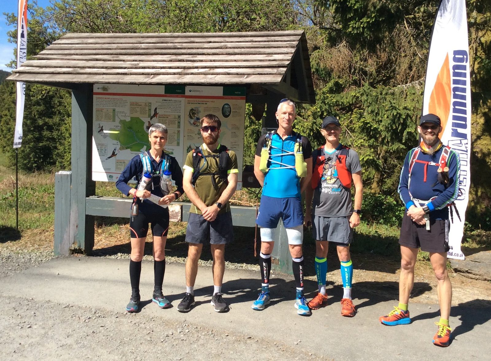 Five runners are standing in front of an information board in a forested area. They are wearing various running gear and hydration packs, under a sunny sky. There is a competitor banner on the right and a gravel path beneath them.