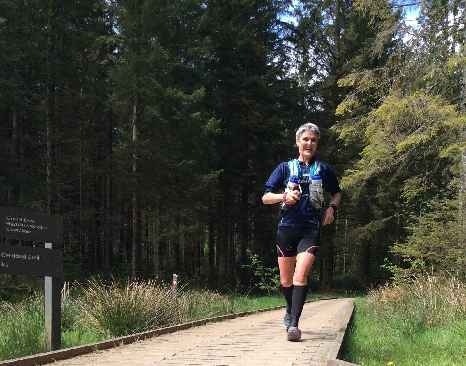 A person with short gray hair runs on a wooden path through a forest, wearing a blue shirt, black shorts, and a hydration pack. The path is surrounded by tall trees and greenery, and a sign with white text is visible to the left of the path.