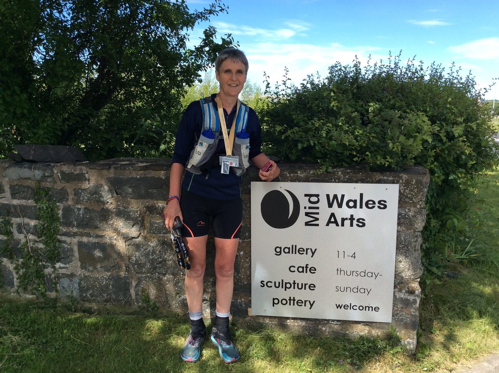 A person wearing athletic gear, holding a pair of sandals, stands beside a stone wall with a sign that reads "Mid Wales Arts: gallery, cafe, sculpture, pottery. 11-4, Thursday-Sunday, welcome." They have medals around their neck and cloudy sky in the background.