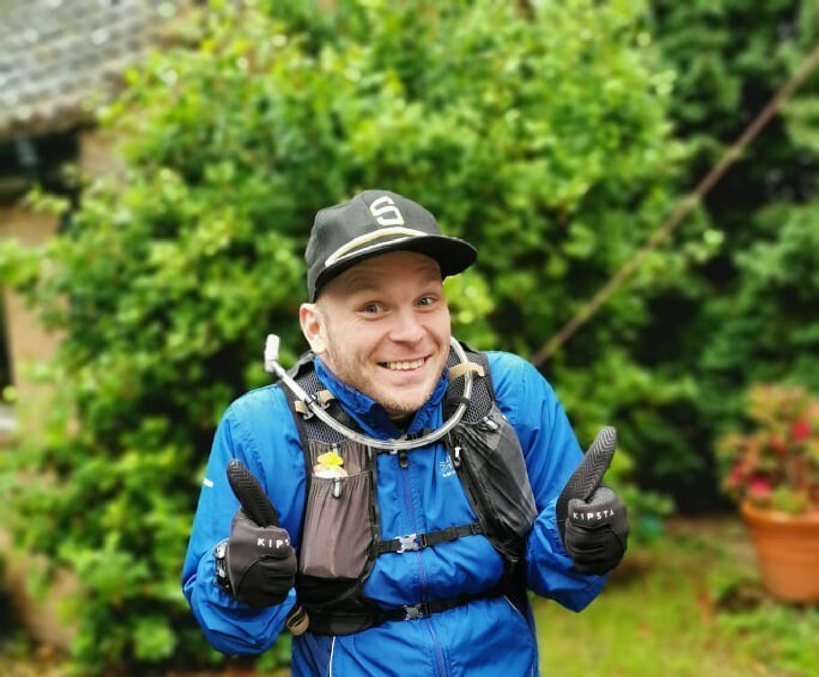 A person in a blue jacket and black cap is smiling and giving a thumbs-up with both hands. They are outdoors in front of green foliage, wearing gloves and a backpack. The sky appears overcast.