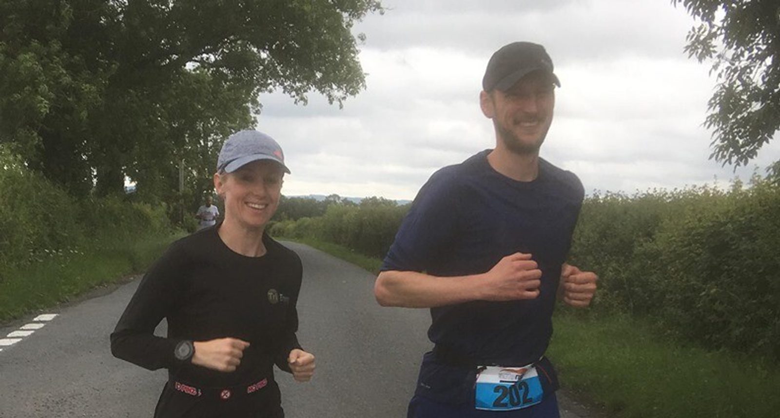 A man and a woman are running outdoors on a country road surrounded by greenery. Both are dressed in athletic wear and smiling. The man wears a blue shirt and black cap, while the woman wears a black shirt and gray cap. They each have a bib number.