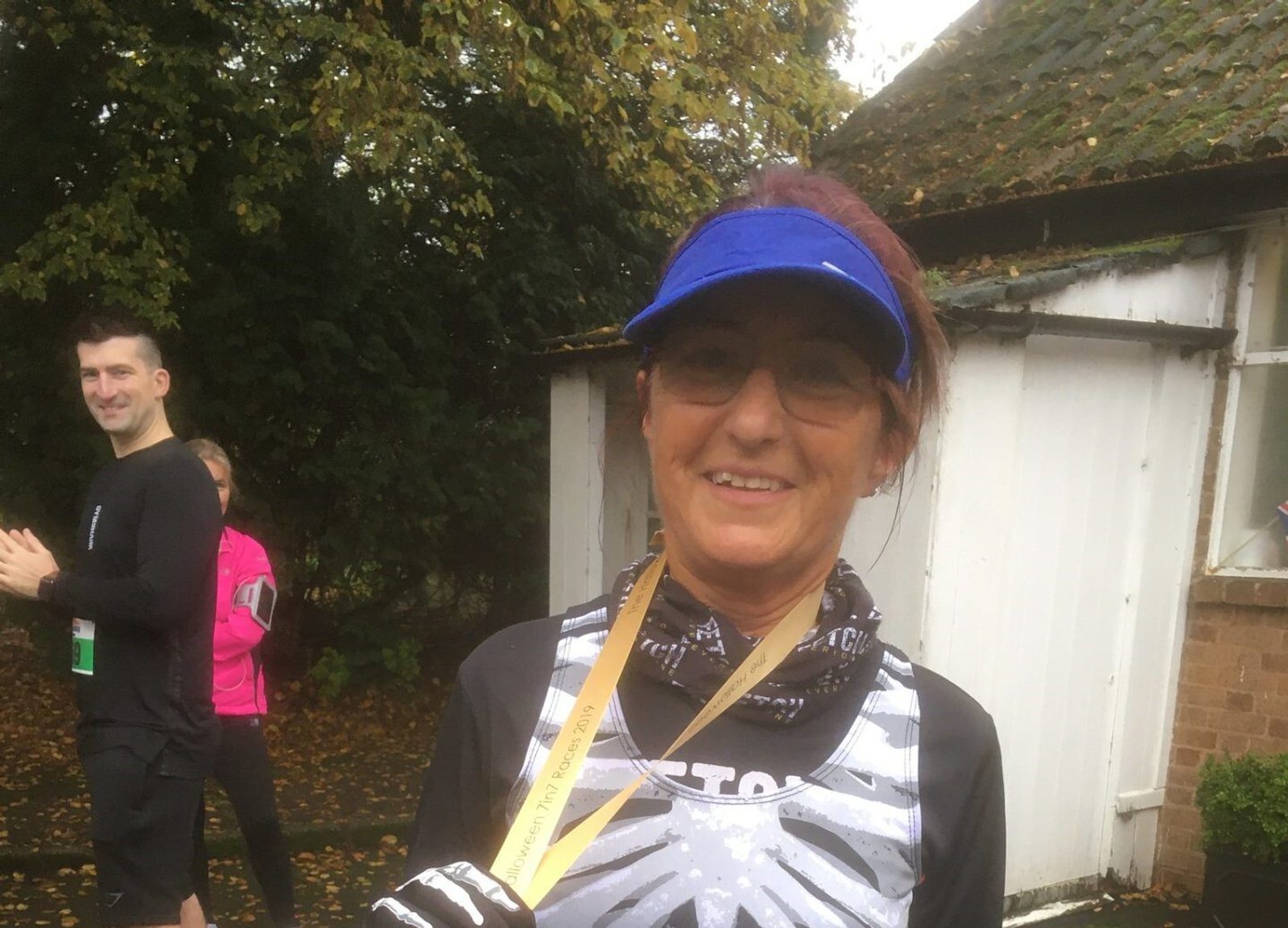 A smiling person wearing a blue visor and black athletic gear with a medal around their neck. They are standing outside with a white shed and trees in the background. Another person in black and one in pink can be seen clapping and holding a drink.