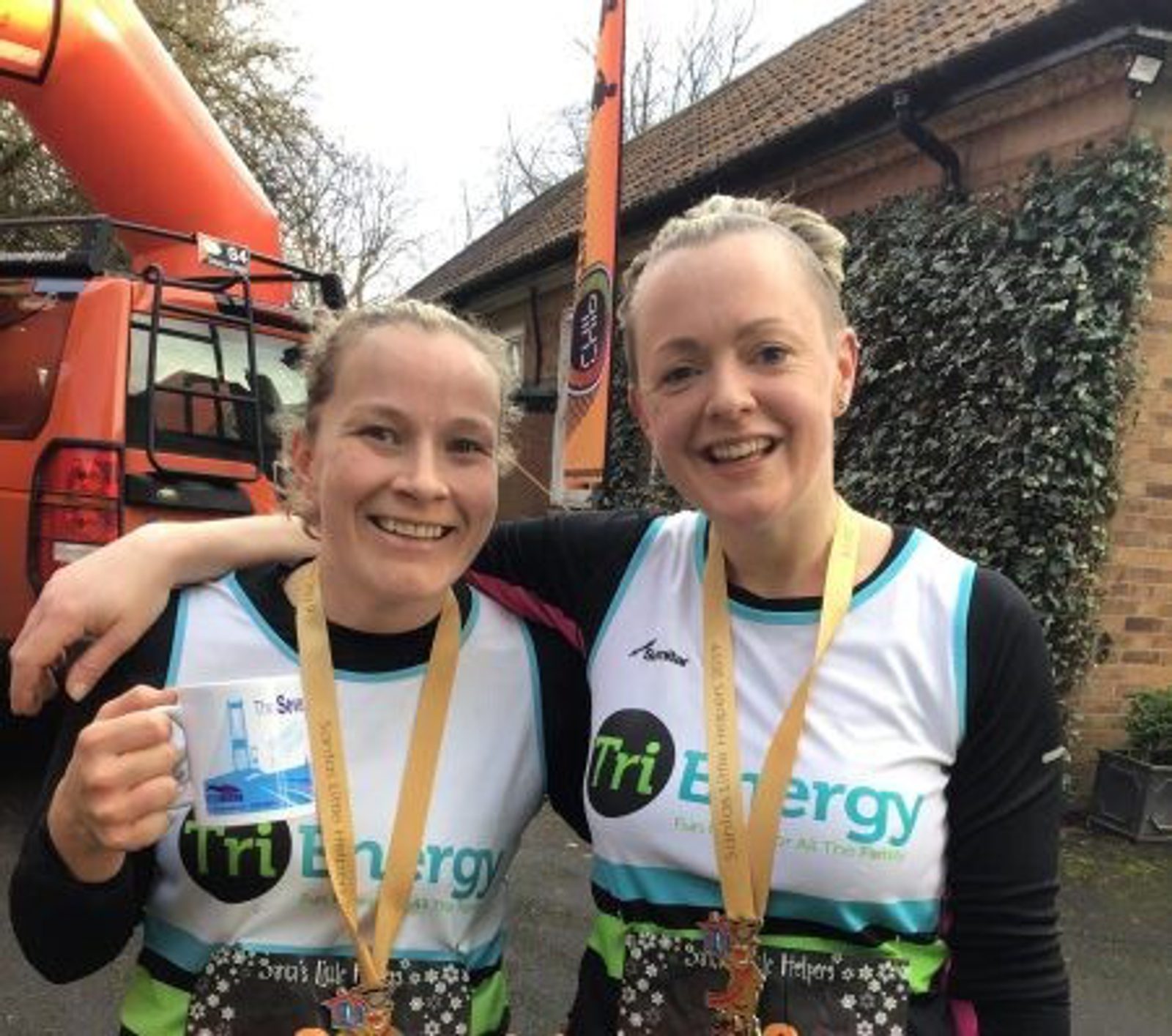 Two smiling women in Tri Energy athletic gear stand side by side with medals around their necks, likely post-race. One holds a mug and the other has her arm around her friend. They are in front of a building with ivy and a red vehicle.