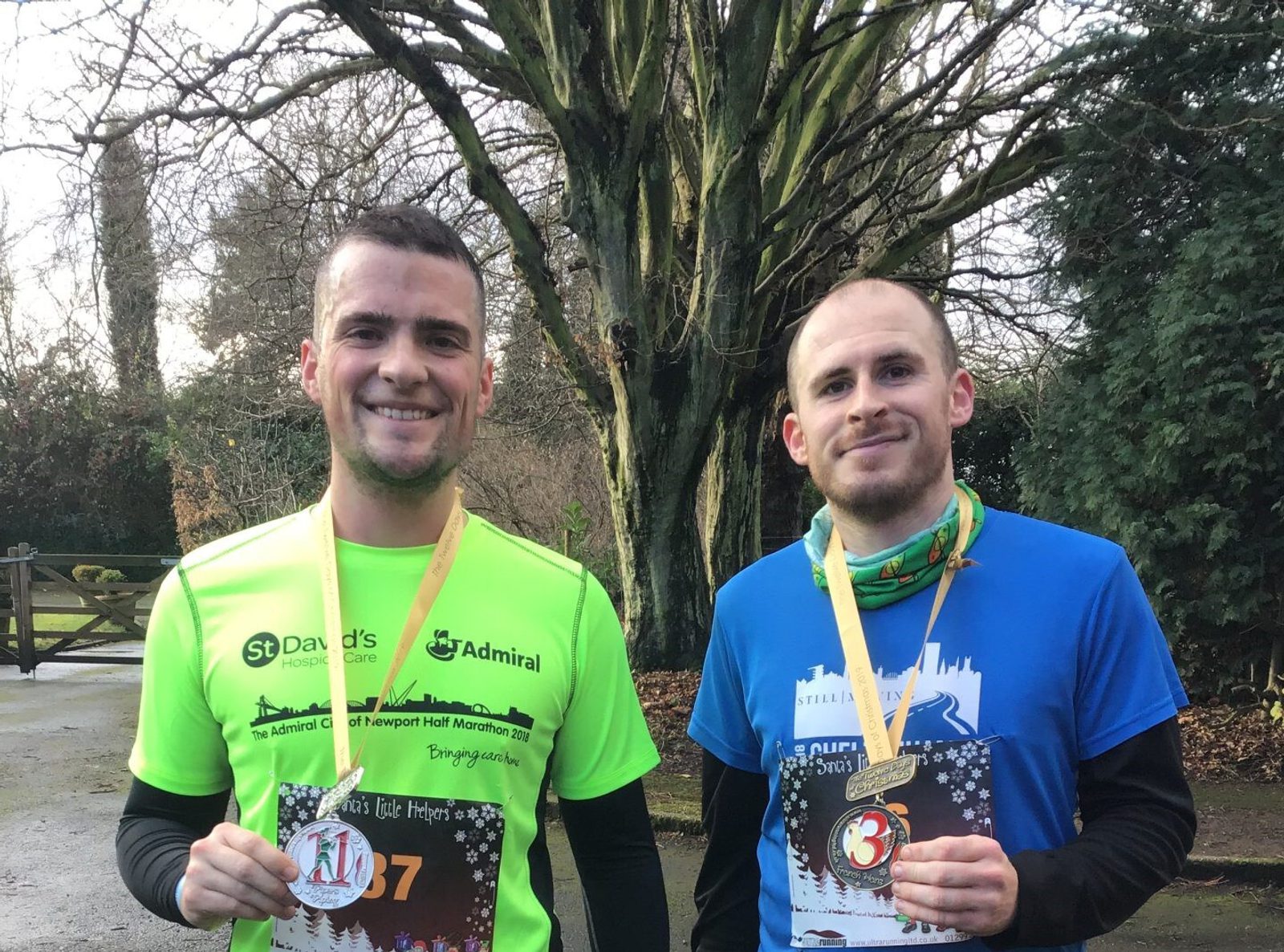 Two men stand outdoors holding medals. The man on the left wears a neon green shirt and holds a medal with a "11" ribbon; the man on the right wears a blue shirt and holds a medal with a "3" ribbon. Both men smile, trees and a wooden gate visible in the background.