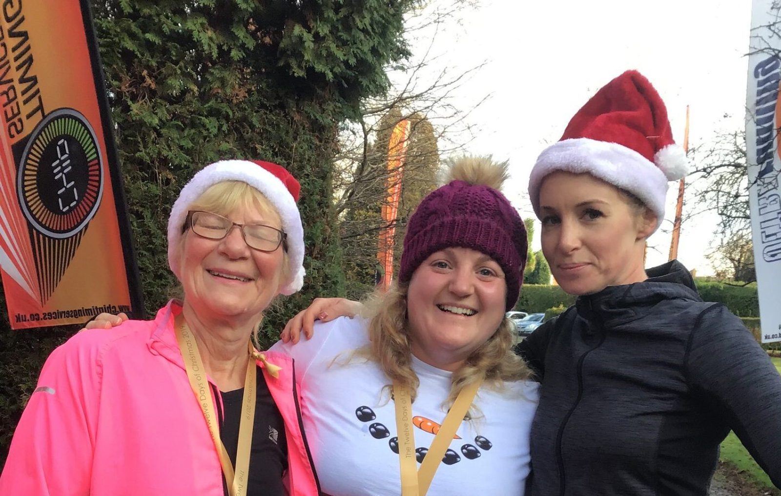 Three women are standing together and smiling, dressed in festive attire with two wearing Santa hats and one wearing a purple knit hat. They appear to be outdoors by a tree. One woman on the left is wearing glasses and a pink jacket. They seem happy and joyful.