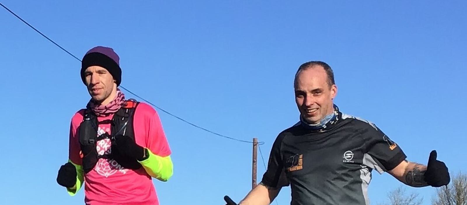 Two men are running outdoors on a clear day. The runner on the left is wearing a pink shirt, a purple hat, and a yellow sleeves, while the runner on the right is in a black shirt. Both are smiling and wearing gloves, with a blue sky in the background.