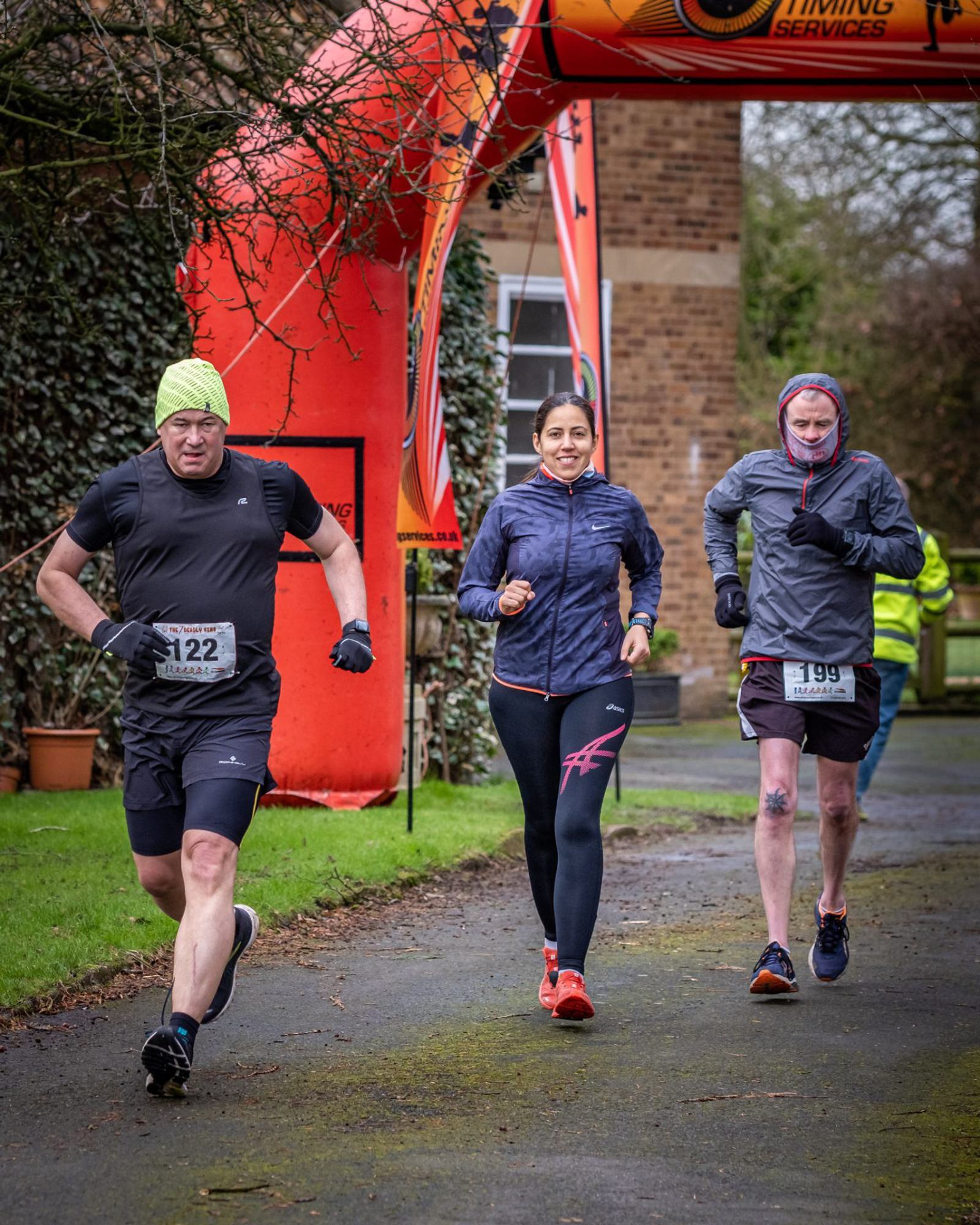 Three runners, two men and one woman, approach the finish line of a race under an orange archway. The man on the left wears a green hat and black shorts. The woman in the center wears a black outfit with red shoes. The man on the right wears a gray jacket and navy shorts.