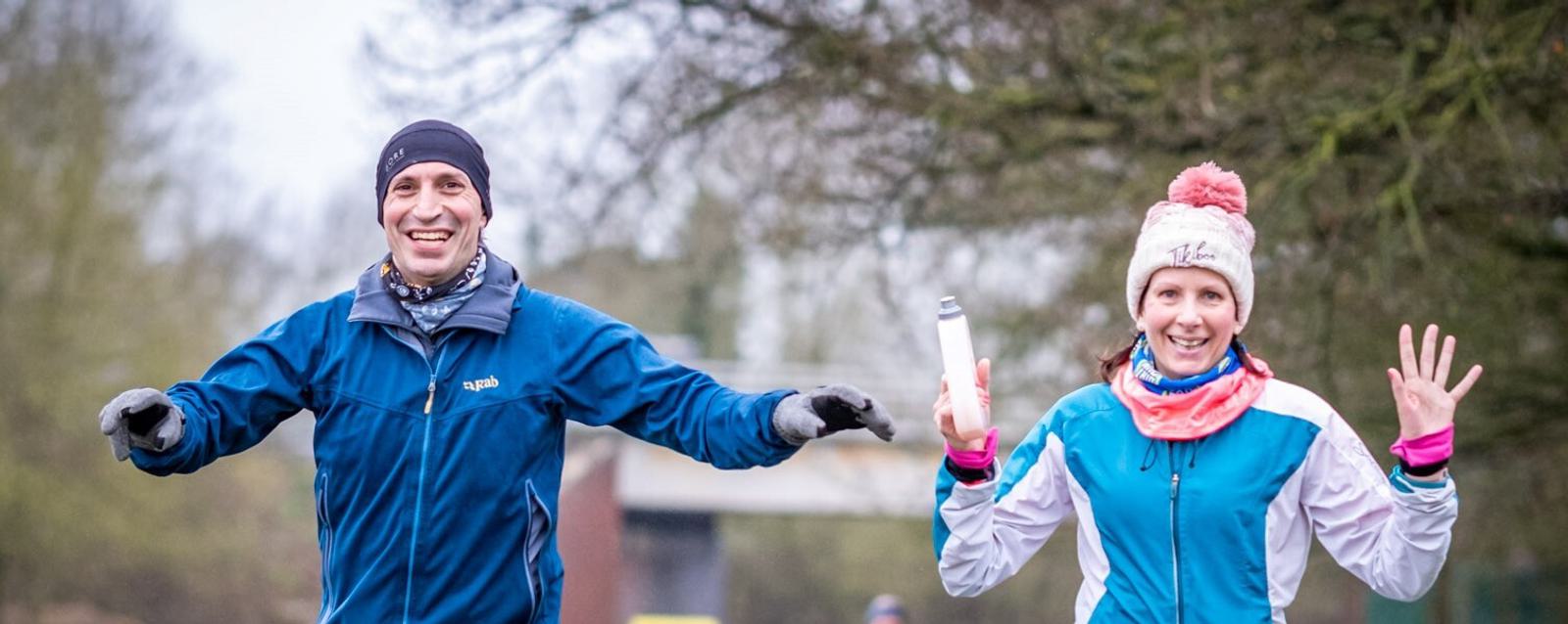 Two people, wearing winter running gear including jackets, gloves, and hats, are running outdoors. They both have cheerful expressions, with the person on the left raising their arms slightly and the person on the right holding a water bottle and raising their right hand.