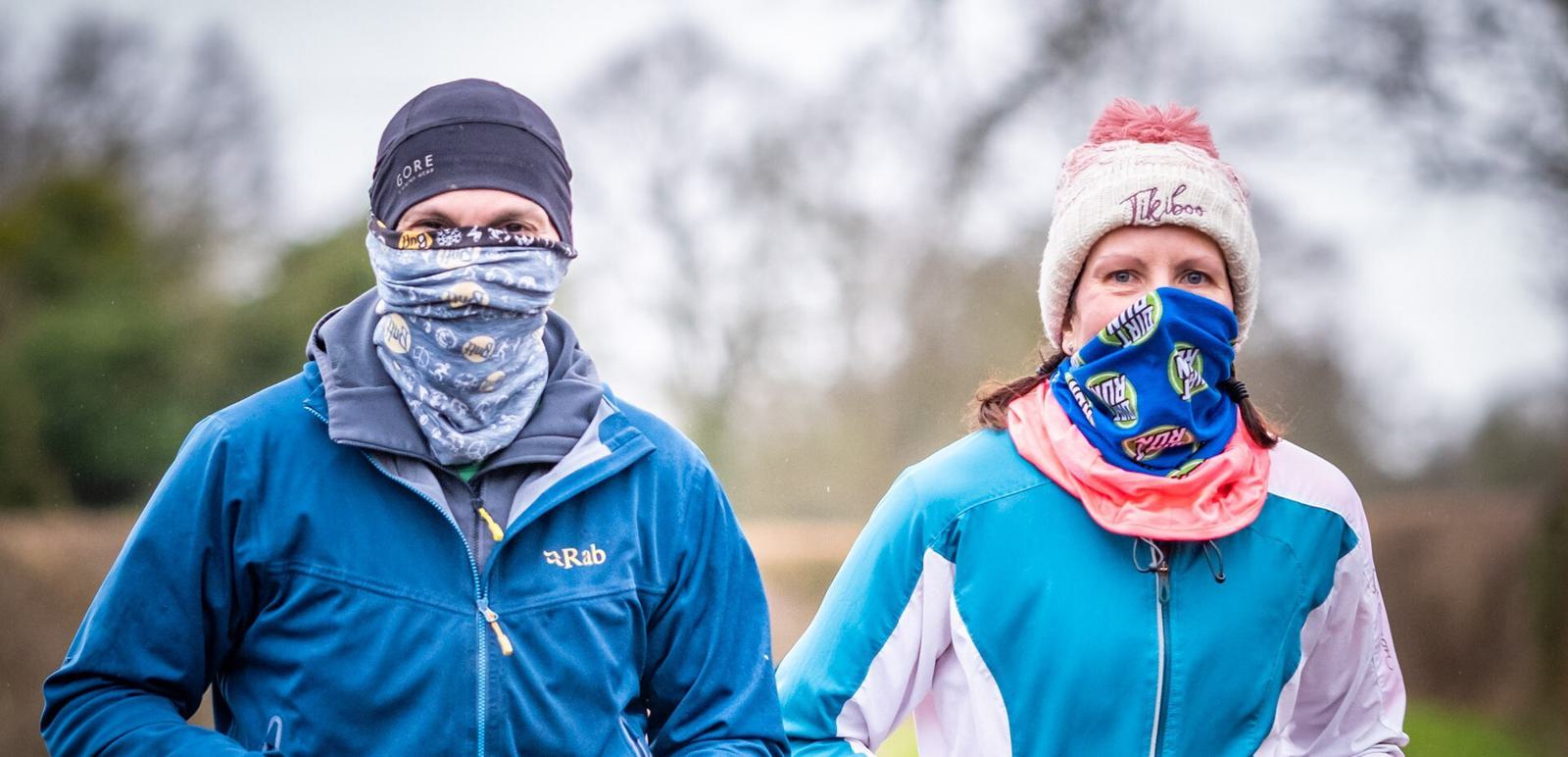 Two individuals dressed in blue jackets and beanies, with bandanas covering their mouths, are jogging outdoors on a cloudy day. The background is blurred, showcasing a natural setting with trees.