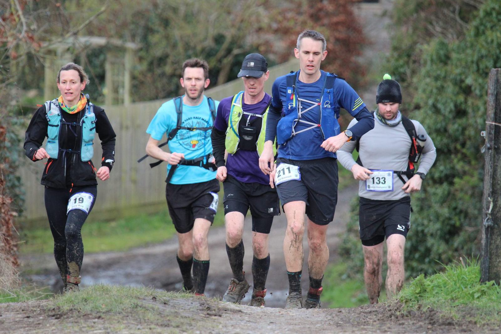 Five runners on a trail, wearing running gear and numbered bibs, are captured mid-race. The path is muddy and surrounded by greenery. The runners appear focused, with some showing signs of exertion.
