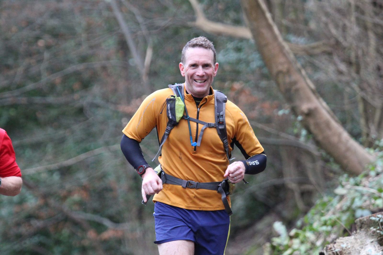 A man in a yellow shirt and blue shorts is running on a forest trail. He is wearing a hydration pack and a watch, and smiling at the camera. The background consists of trees and greenery. Another person in a red shirt is partially visible to his left.
