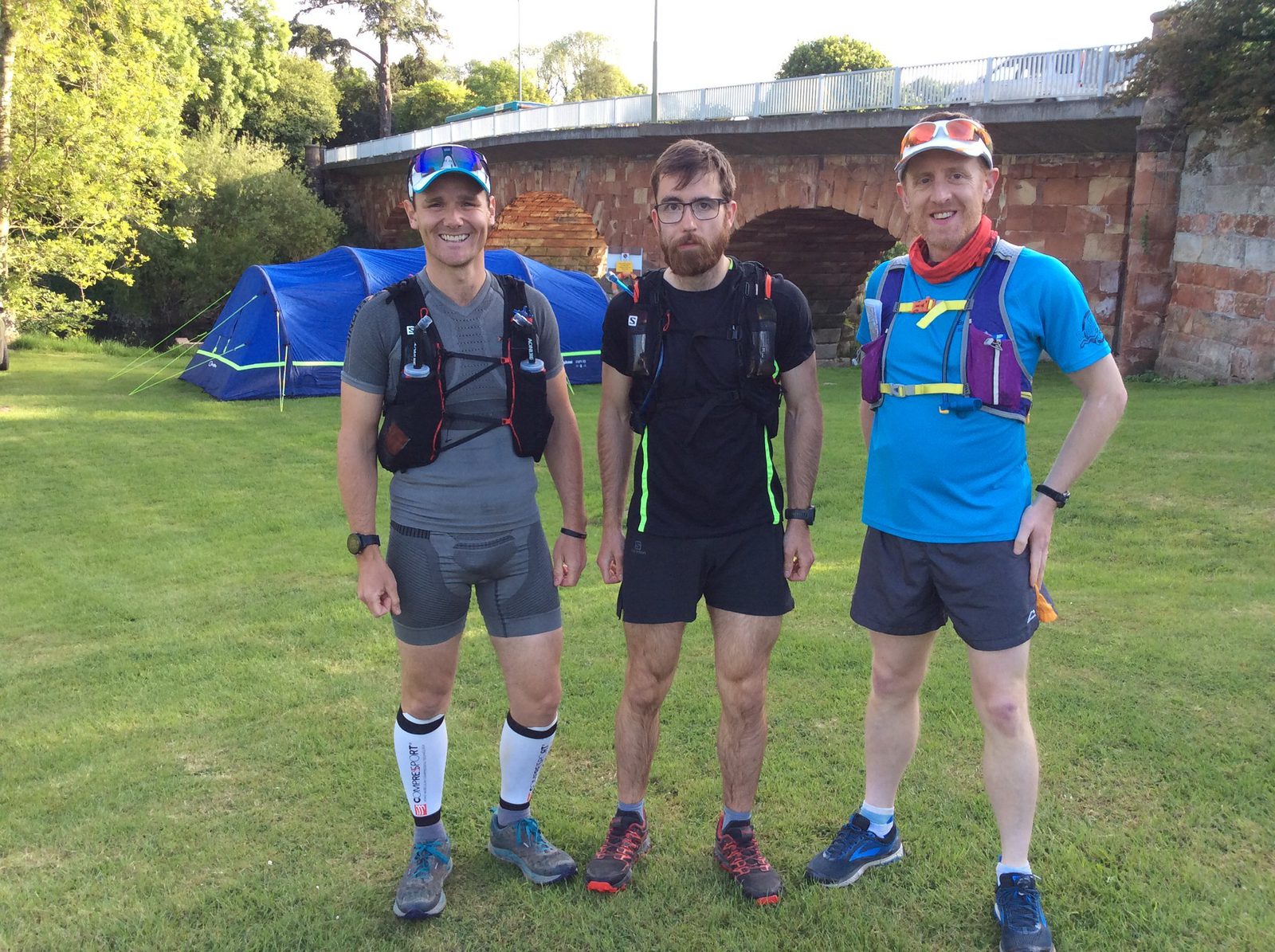 Three men wearing athletic gear and backpacks stand on grass near a bridge and a blue tent. They are smiling and seemingly prepared for a sporting activity, possibly trail running or hiking, with lush greenery and bright sunlight in the background.