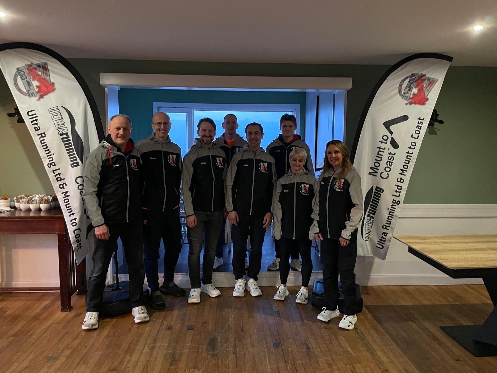 A group of eight people is standing indoors, all dressed in matching black and grey outfits with a logo on the left chest. Two large banners on either side of the group also display the same logo, with the text "Ultra Running Ltd Mount to Coast" written on them.