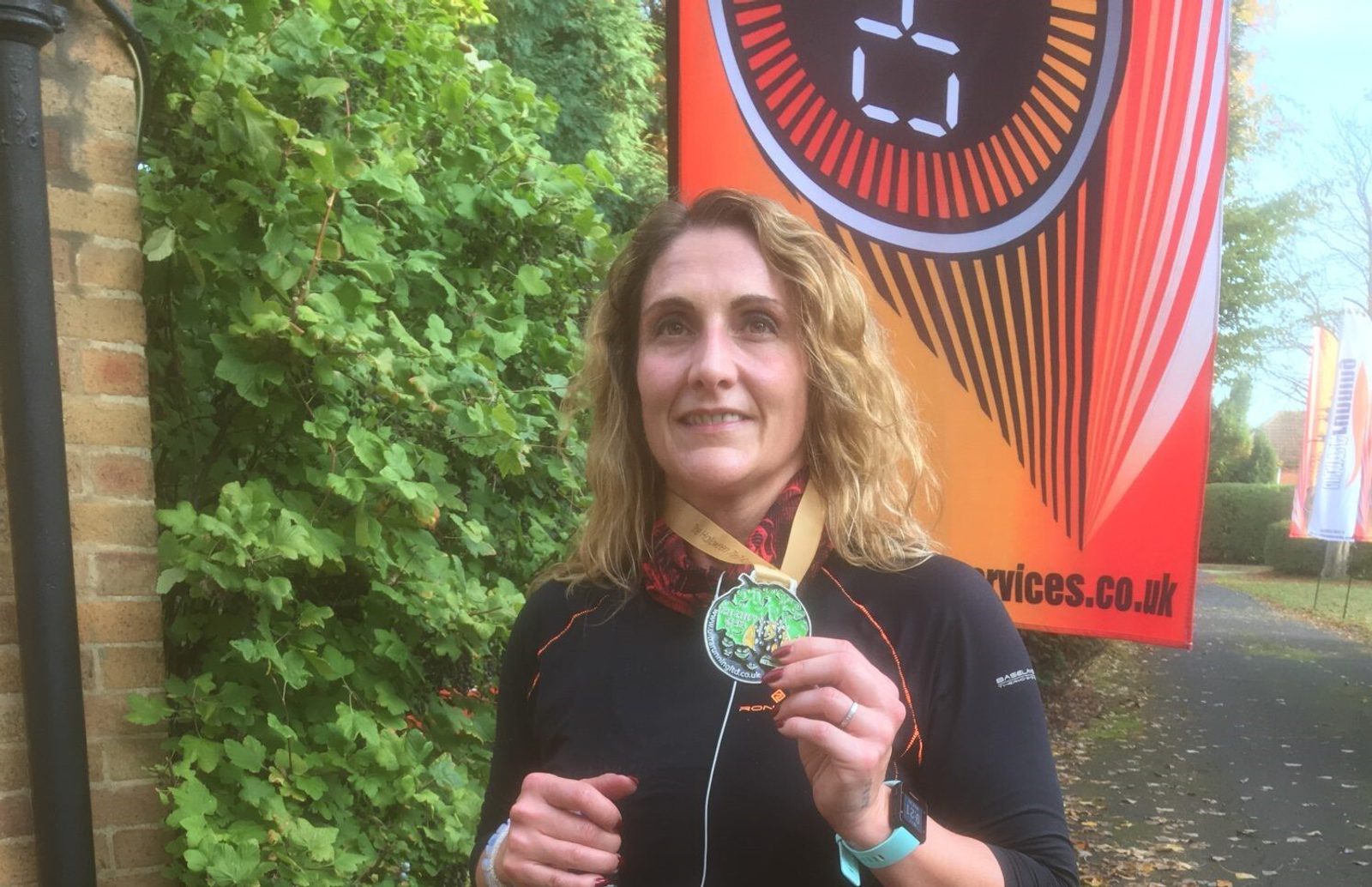 A woman with curly blonde hair is standing outdoors, smiling and holding a medal around her neck. She is wearing a black sports top. Behind her is a banner with the number 2 on it and some greenery. The scene suggests a recent achievement, possibly in a race.