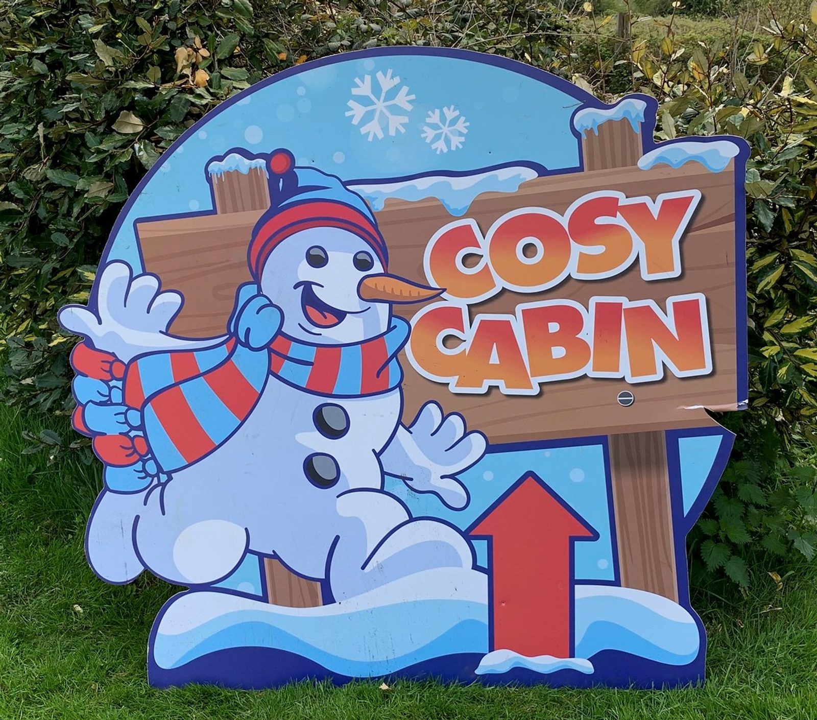A colorful sign featuring a cheerful snowman wearing a red hat and striped scarf points toward the "Cosy Cabin." The sign has a backdrop of snowflakes and a wooden arrow directing viewers to the cabin, standing in a grassy area with bushes in the background.