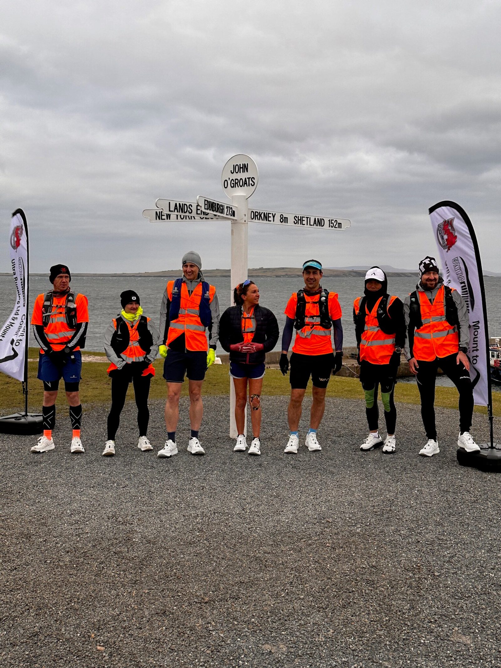 A group of seven people wearing bright orange vests and athletic wear are standing in front of the John O'Groats signpost. They appear ready for a run, and the backdrop shows a grey, cloudy sky over the ocean. Two flags with logos stand on either side of the group.