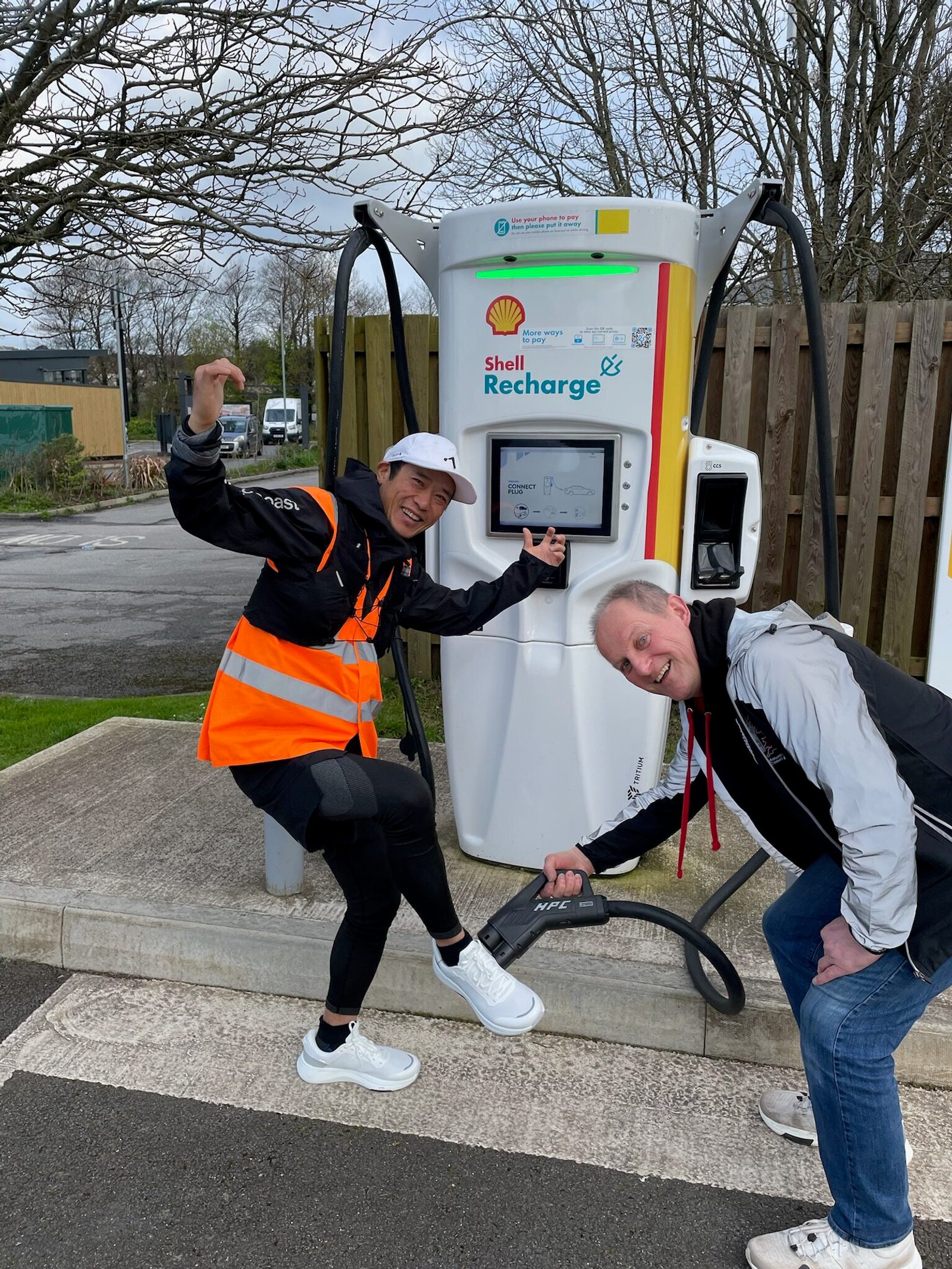 Two smiling people, one in an orange safety vest and the other in a black and gray jacket, happily demonstrate a Shell Recharge electric vehicle charging station. The person in the jacket is holding the charging nozzle, while the other points at the display screen. Trees and a wooden fence are visible in the background.