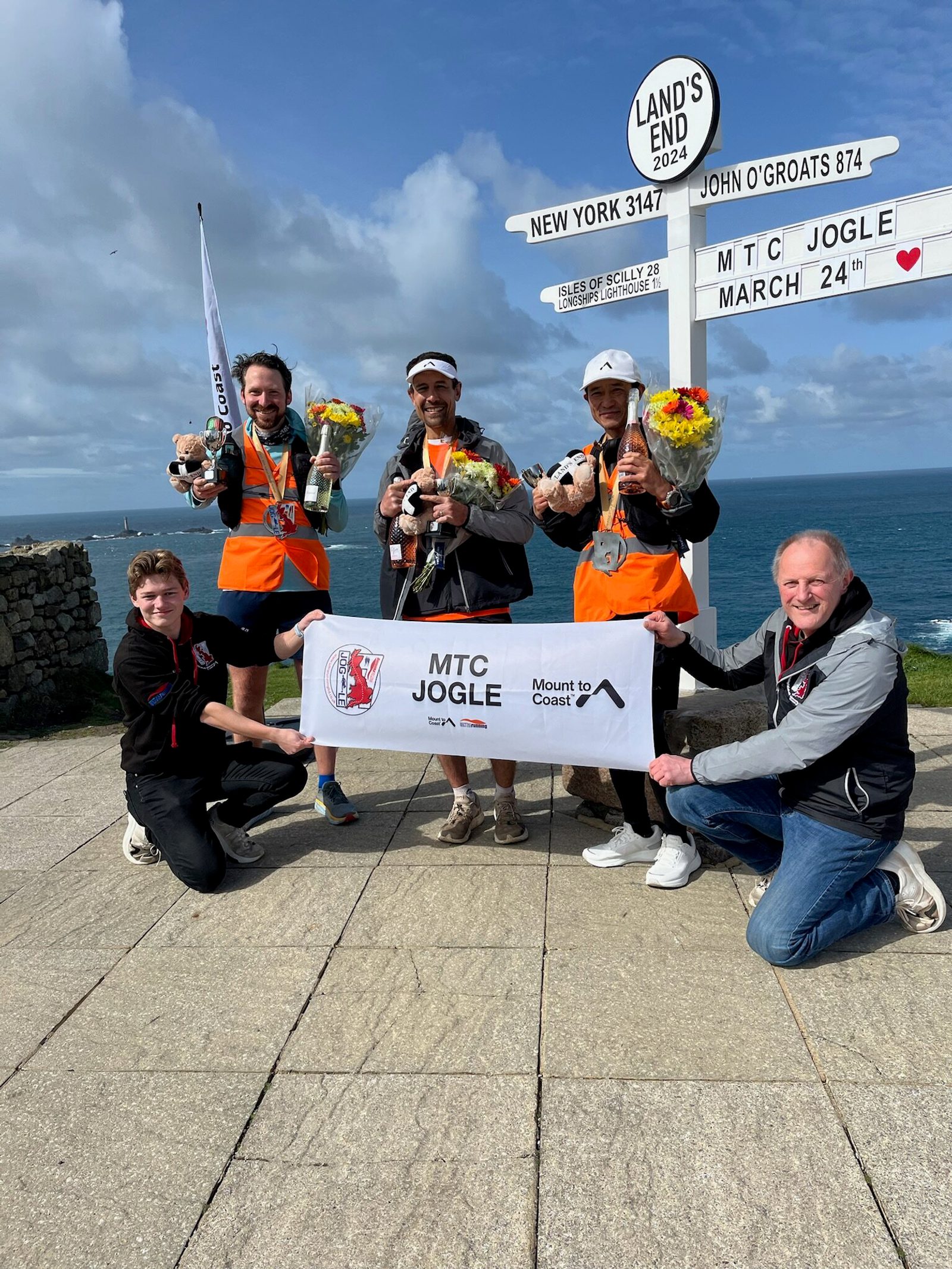 A group of five people stand and kneel near a signpost at Land's End. Three are wearing orange vests and holding stuffed animals and flowers. They hold a banner reading "MTC JOGLE Mount to Coast." The background features the sea and a cloudy sky.