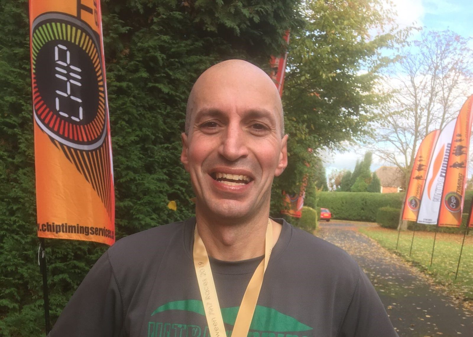 A smiling man with a shaved head is wearing a medal around his neck and a gray shirt with a green logo. He stands on a paved path lined with trees. Behind him are several orange flags with text on them, indicating a race or event setting.