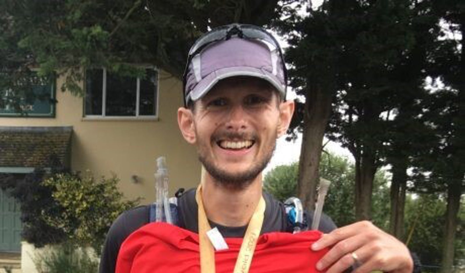 A smiling person wearing a cap and a black shirt holds up a red shirt. They are also wearing a medal around their neck. Trees and a building with a green door are visible in the background.