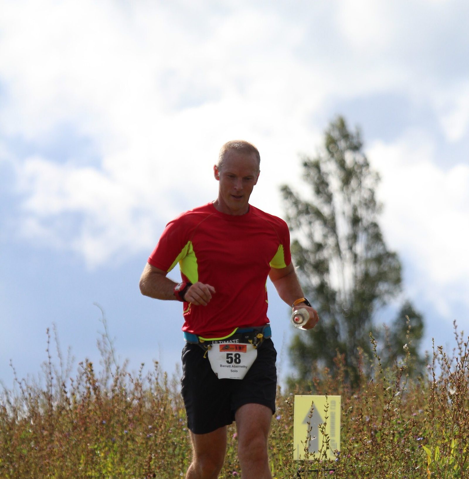 A runner in a red and yellow shirt with the bib number 58 is jogging through a grassy area with a cloudy sky in the background. A directional sign with an arrow is visible on the right side of the image, indicating the race route.