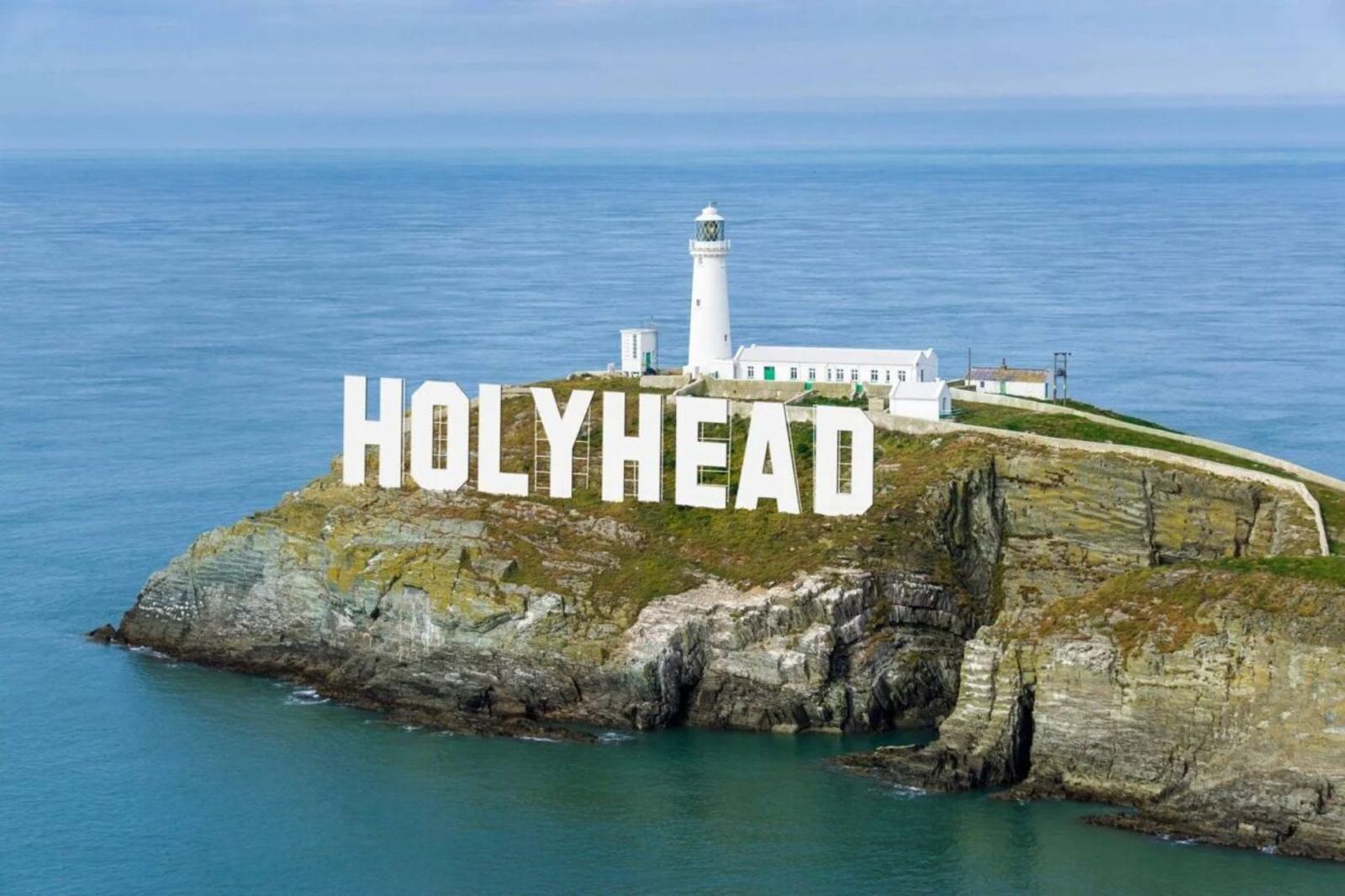 White lighthouse and large "HOLYHEAD" letters on a rocky cliff by the sea. Rugged coastline with grassy patches and a clear blue sky above. A narrow path leads up to the lighthouse.