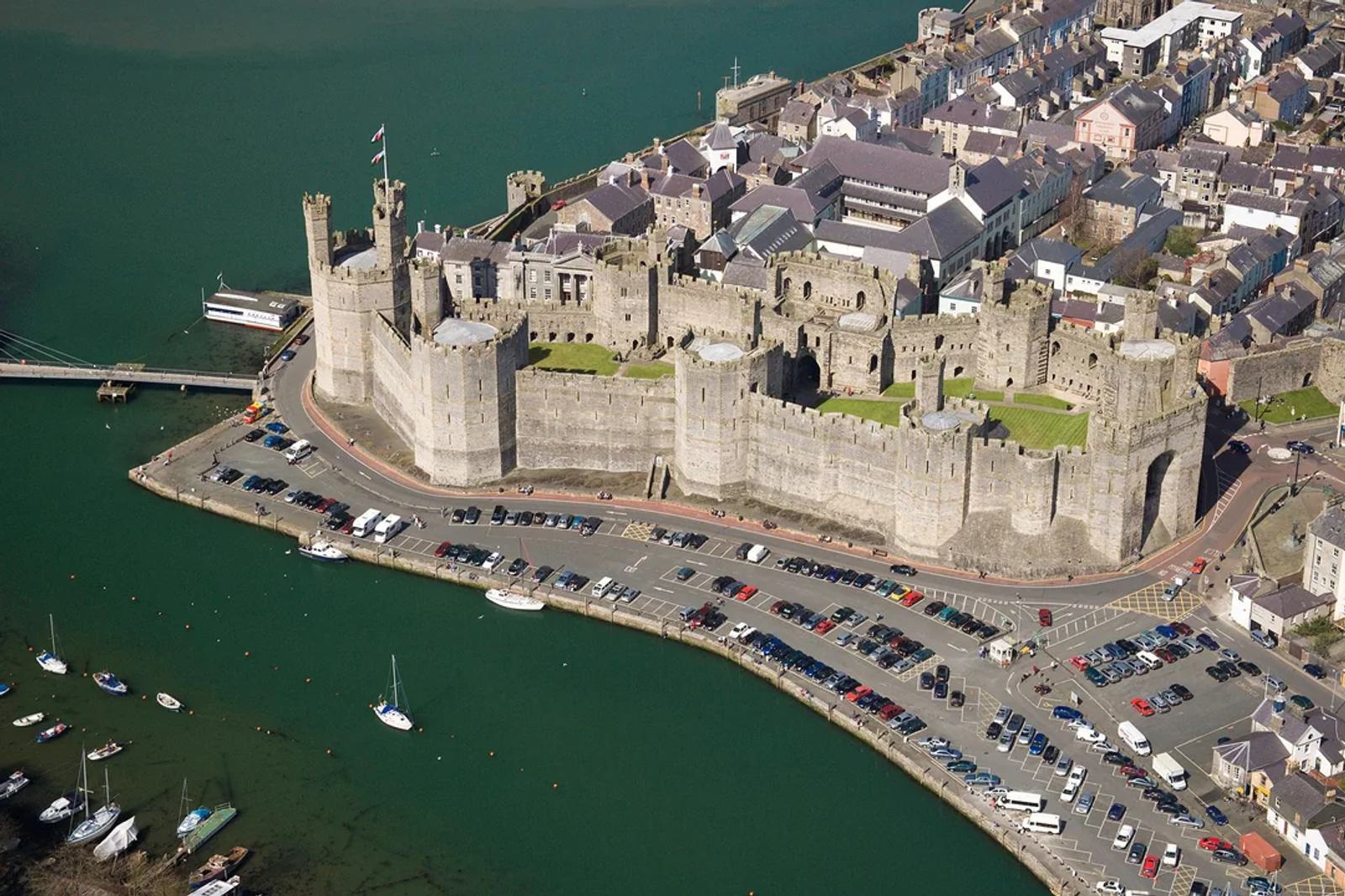Aerial view of a medieval castle with multiple towers, surrounded by a waterway. The castle is adjacent to a town with closely packed buildings. Cars are parked along the waterfront, which hosts small boats. A bridge connects to the opposite shore.