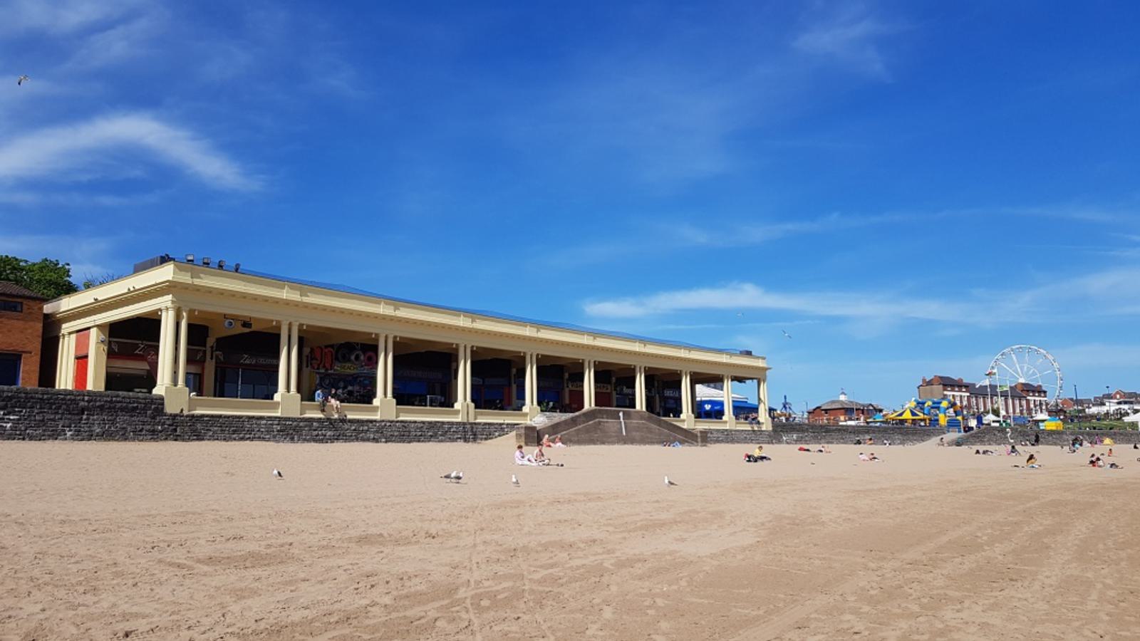 A sandy beach with scattered people sunbathing and seagulls nearby. In the background, there's a yellow building with stone steps and a Ferris wheel and colorful structures, possibly a fairground, under a clear blue sky.