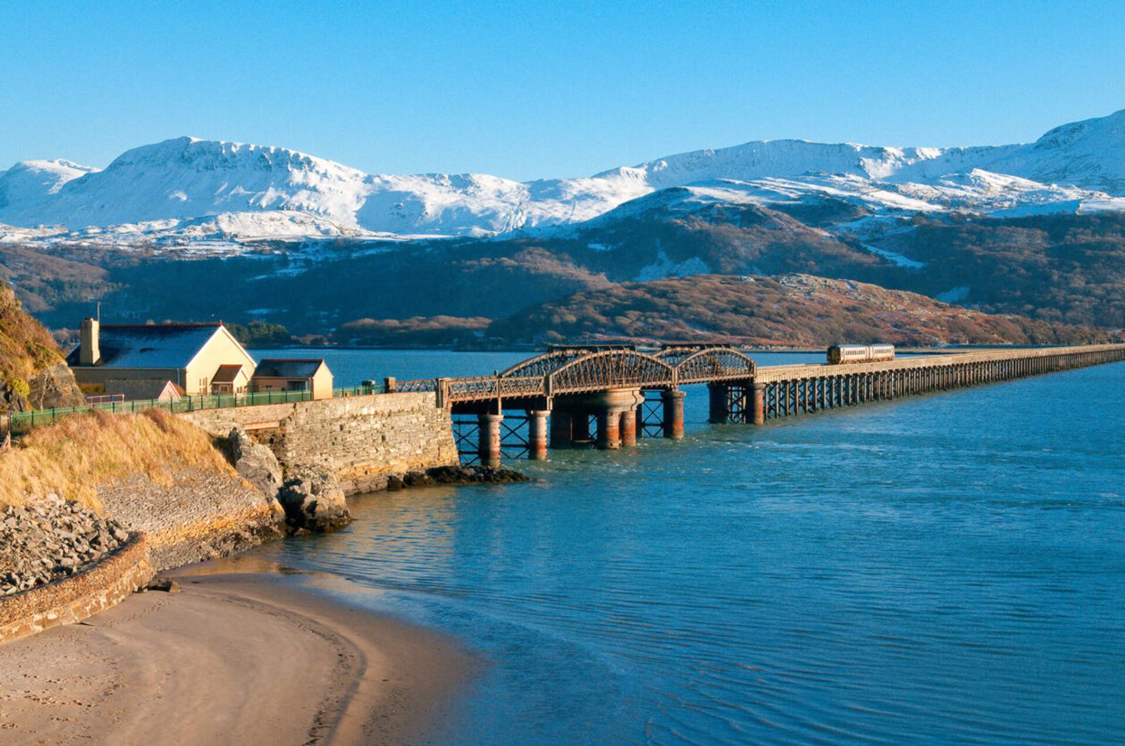 A long, arched railway bridge stretches across a calm blue body of water, leading towards snow-capped mountains. A small sandy beach and houses are in the foreground, with bright, clear skies overhead.